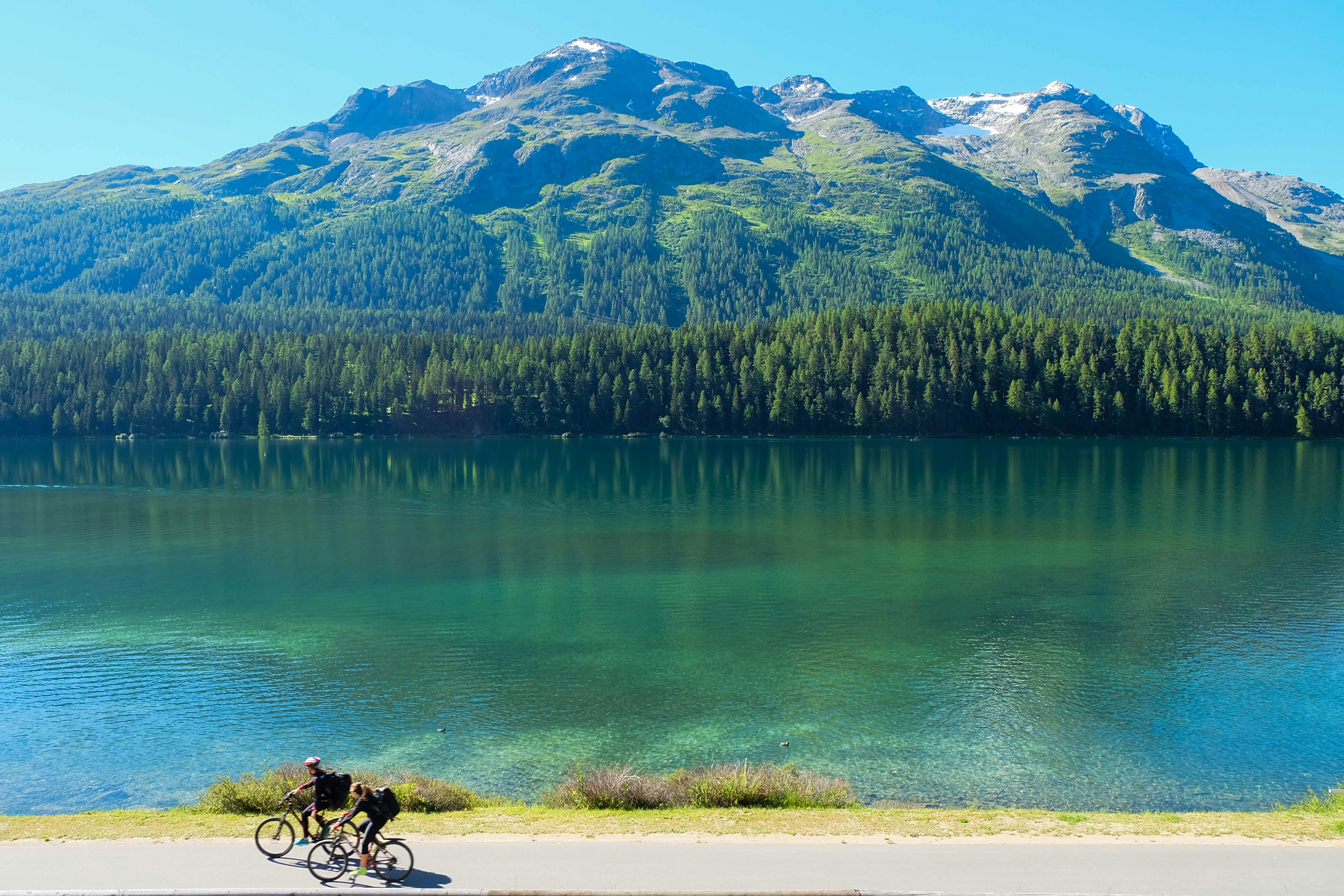 Two cyclists bike along a road on the banks of a large lake, near Lugano in Switzerland. The lake's water is a beautiful blue and flanked by green forest. 