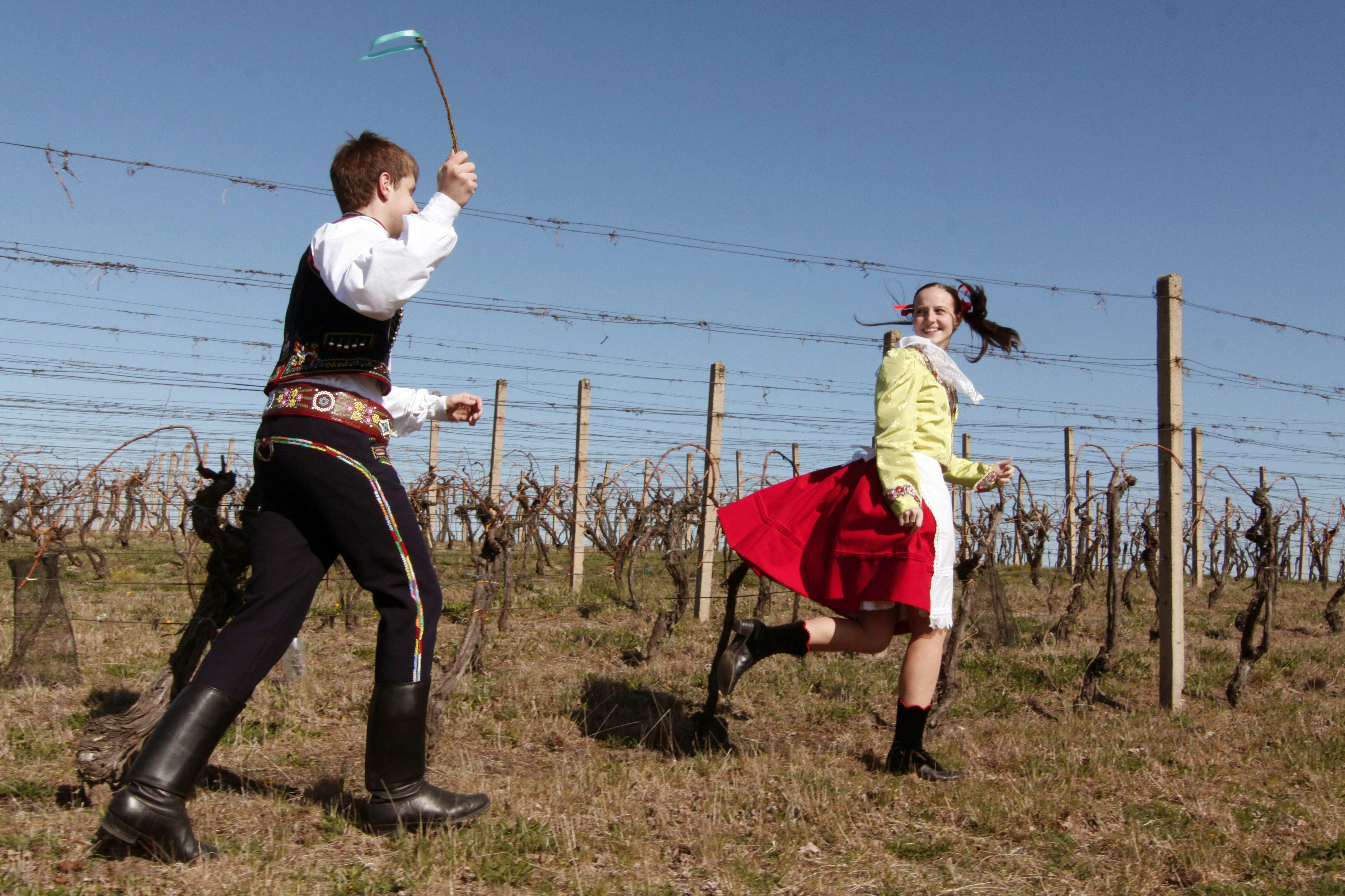 A young man dressed in traditional Czech costume and holding a braided whip is running towards a girl; the girl is running away, laughing.