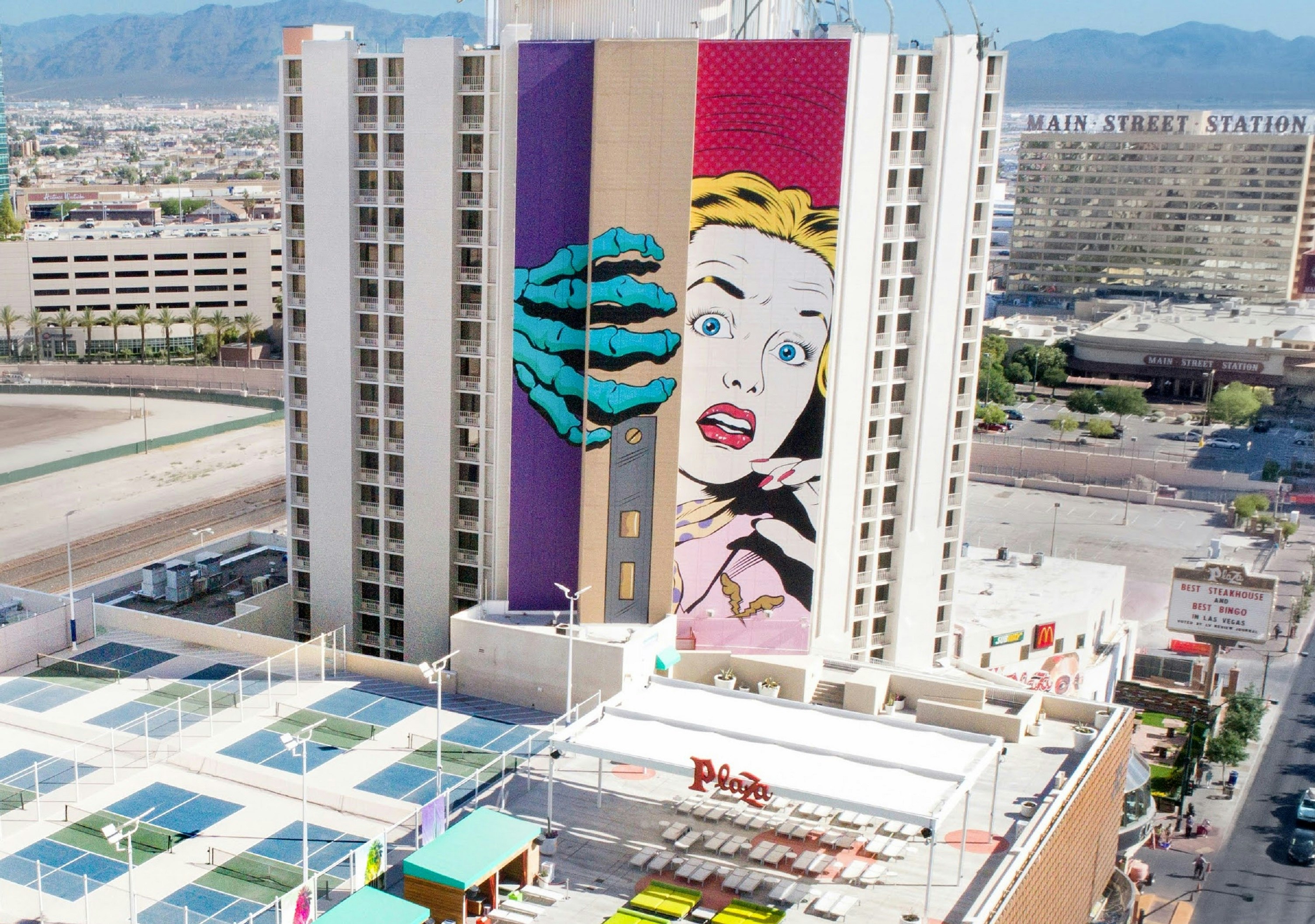 A wide shot of a 21-story mural on the side of the Plaza Hotel & Casino in Las Vegas; it shows a colorful, comic-book-style image of a scared woman and a skeleton's hand opening a door. 