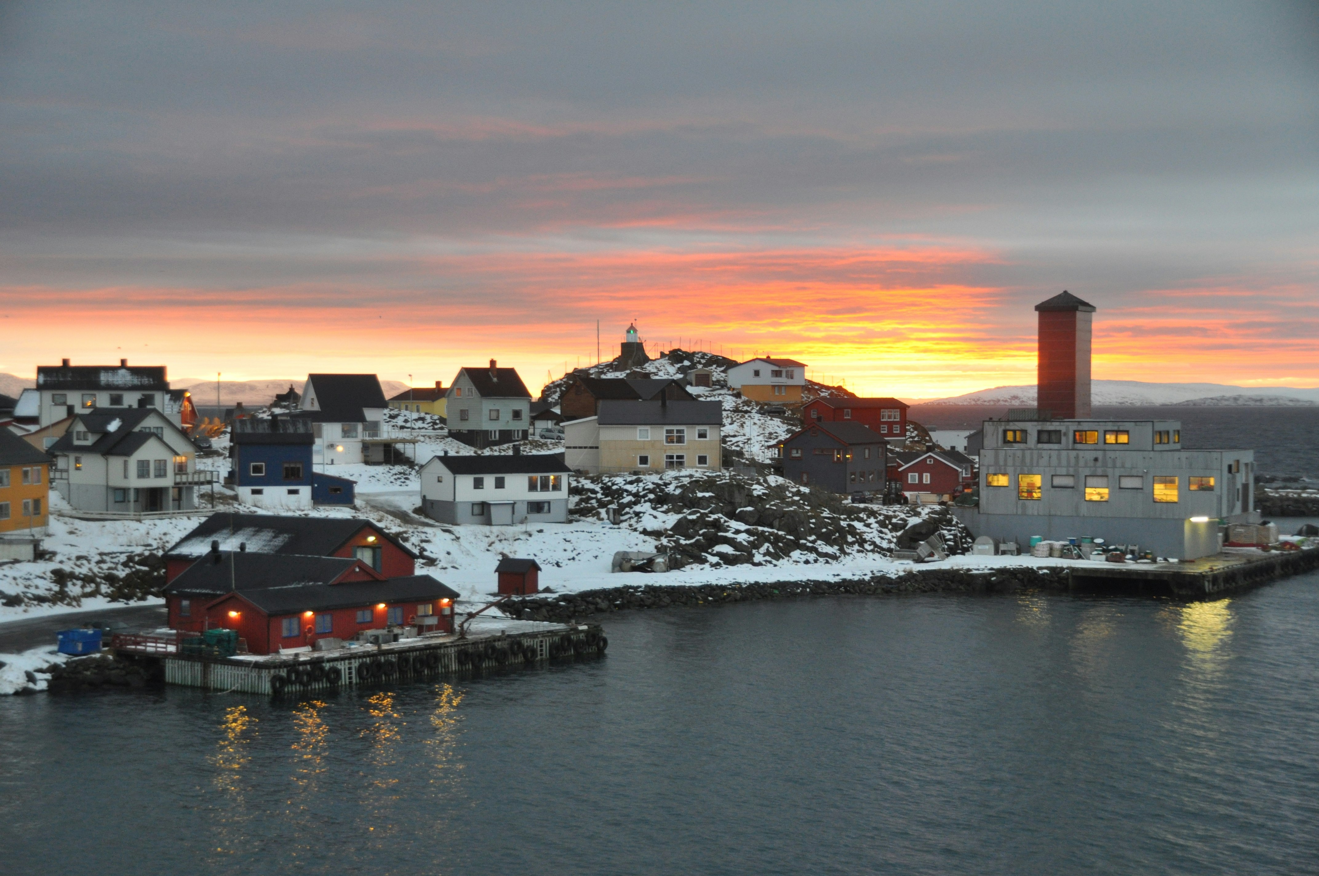 The port of a small coastal town in Norway. It's approaching sunset, and the sky near the horizon has a blazing orange glow, while there is sno dusting the buildings and ground in the town.
