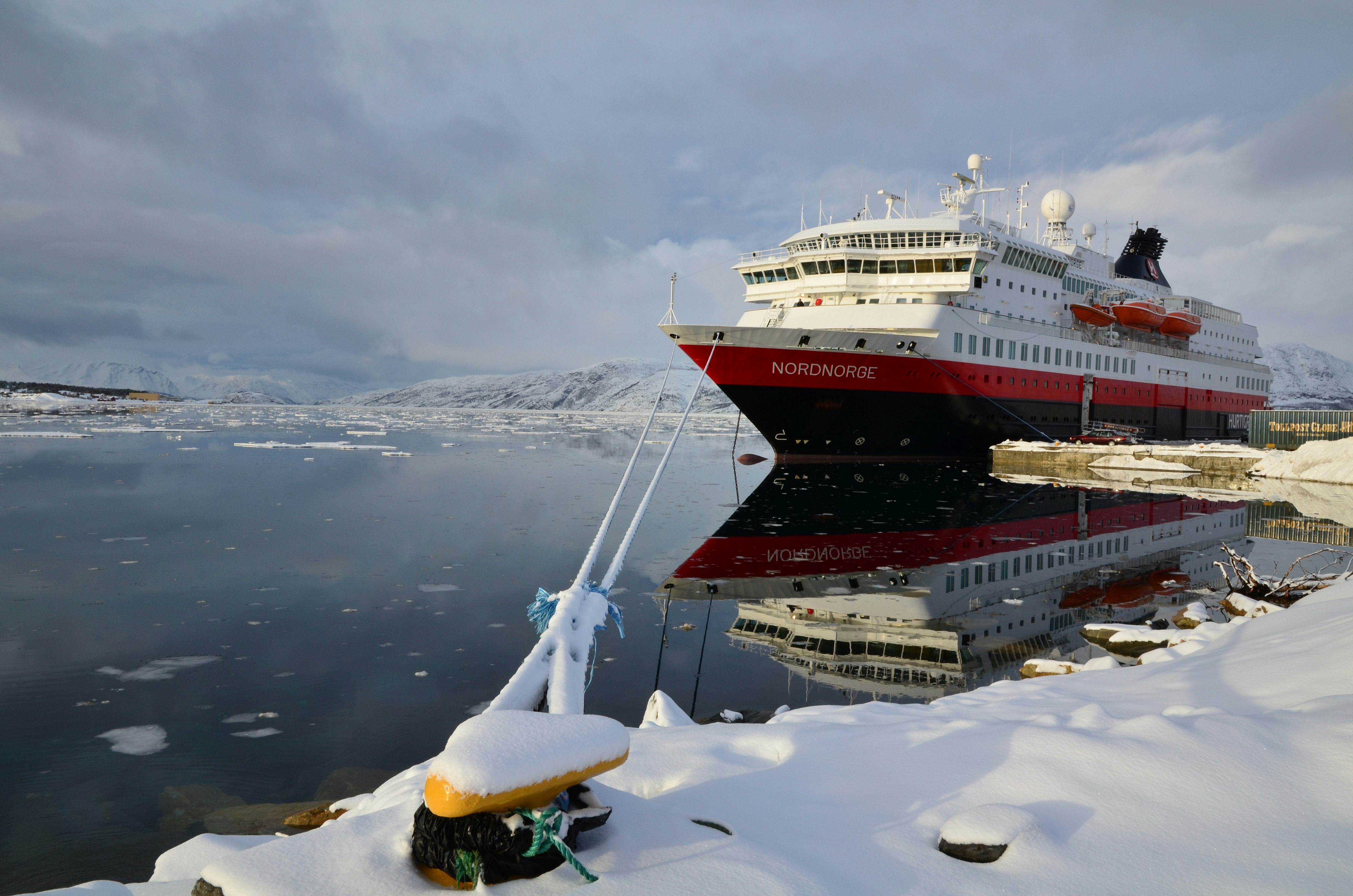 A large passenger ferry named the Nordnorge is docked at a port in winter. It's a snowy scene, and patches of ice can be seen in the water.