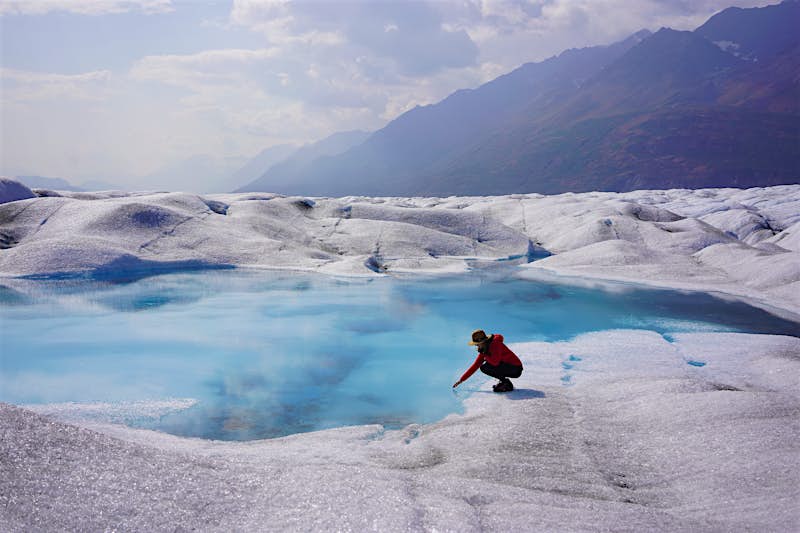 a woman crouches low over a brilliant blue lake on the top of a glacier with mountains in the background