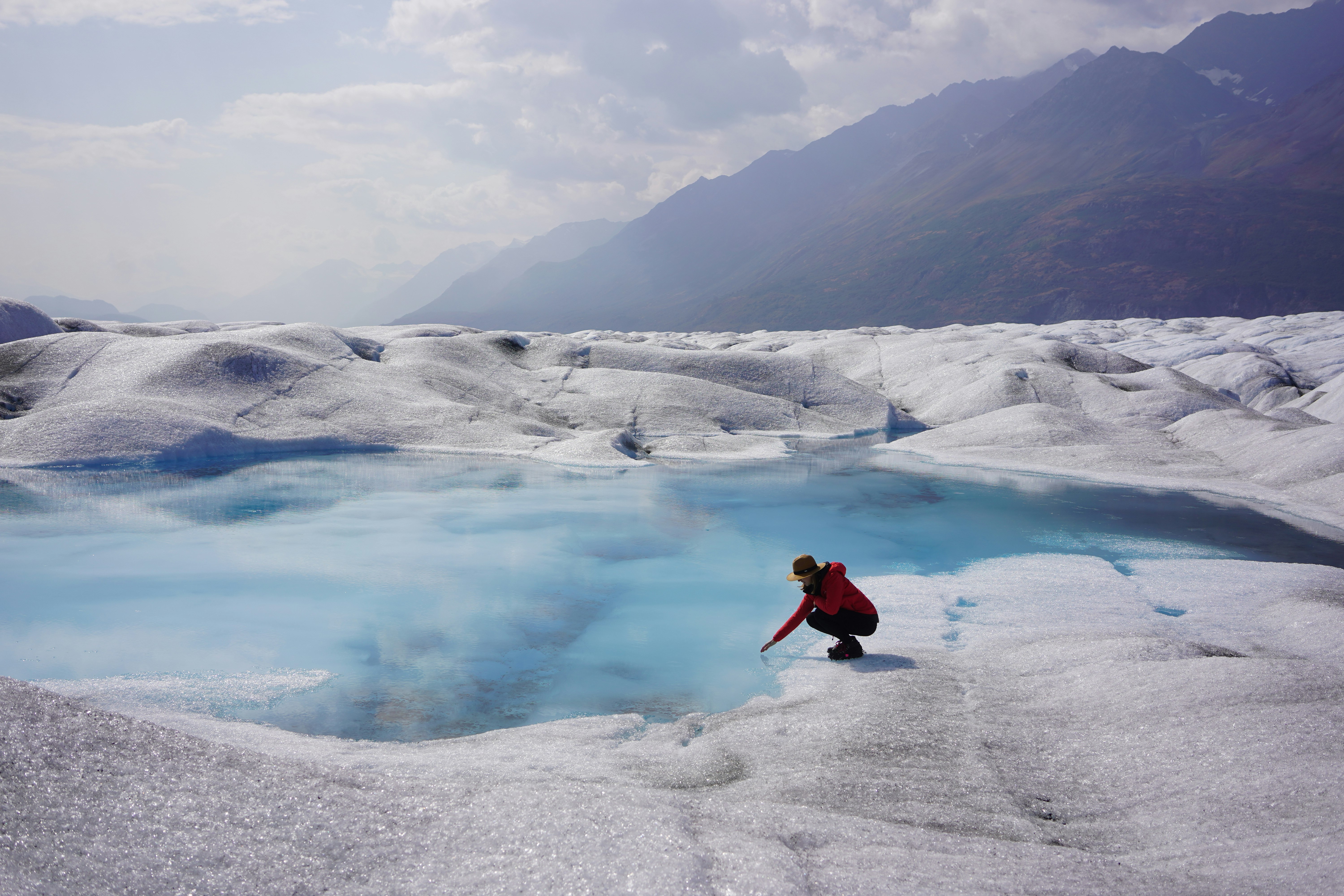 a woman crouches low over a brilliant blue lake on the top of a glacier with mountains in the background