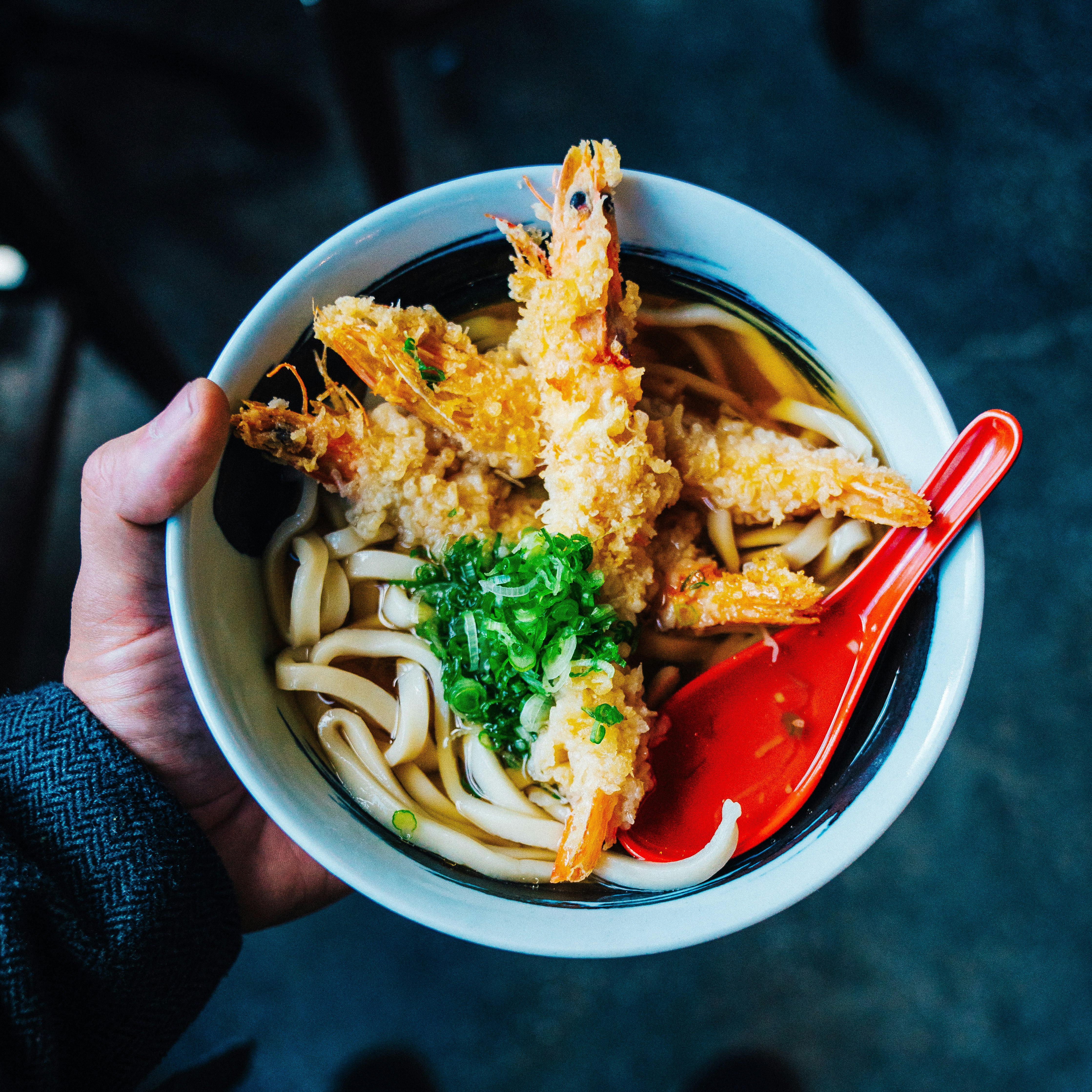 A hand holds a bowl with noodles, broth and fried prawns