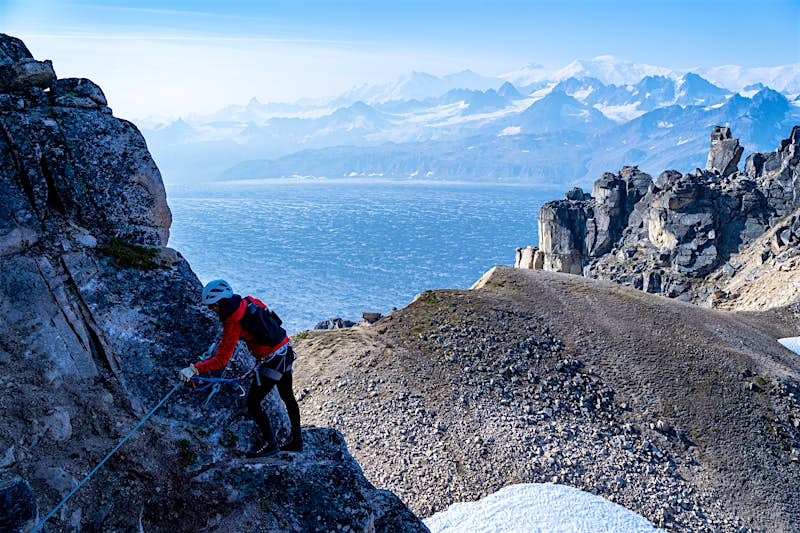 A woman picks her way along a rock wall with a mountain view and ocean in the background