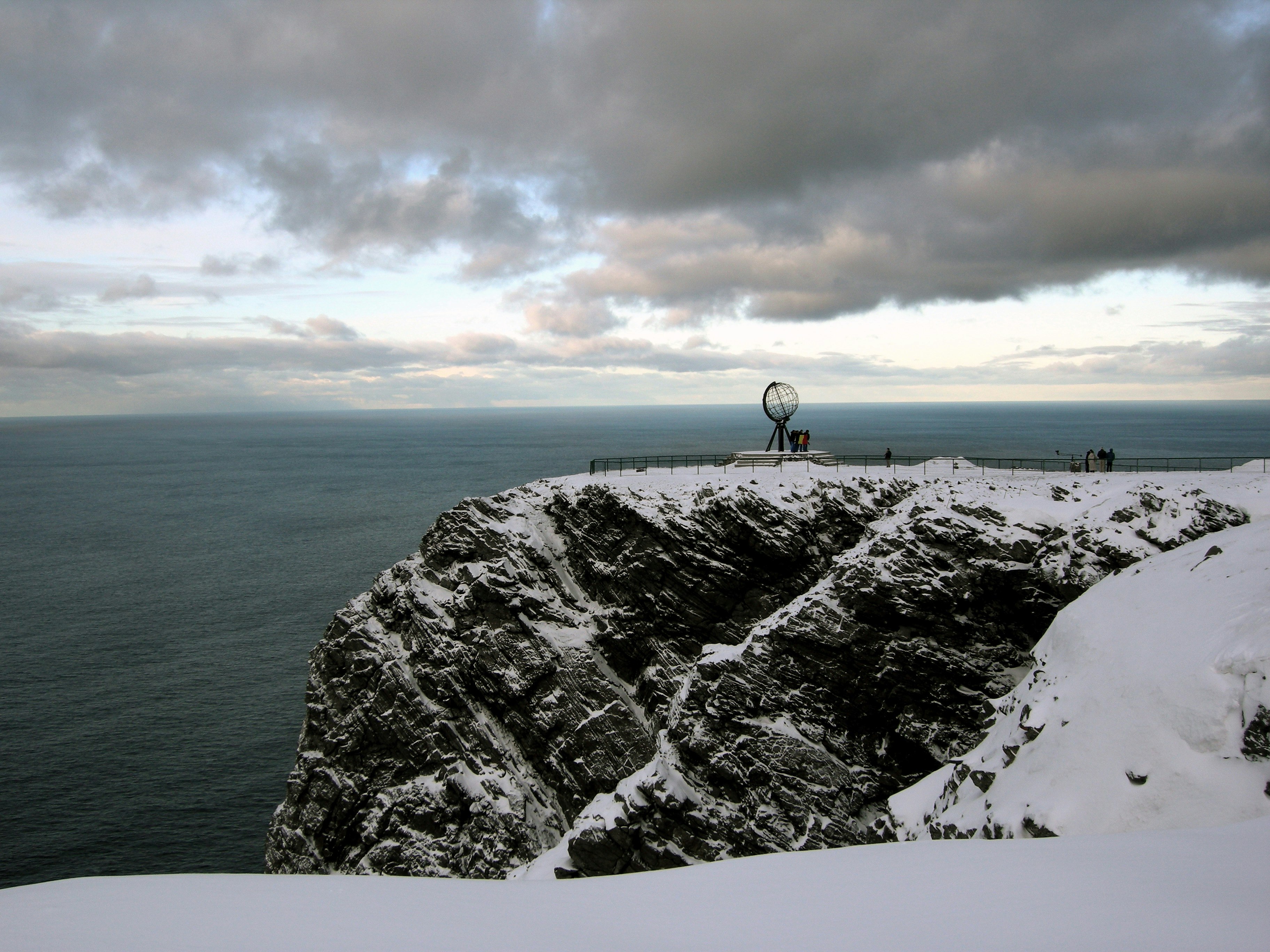 A scene of a snow-covered, rocky cliff-face, with dark grey-blue sea visible below and beyond. There is a globe-shaped monument on the cliff, and a few people can be seen around it, and looking out at the view.