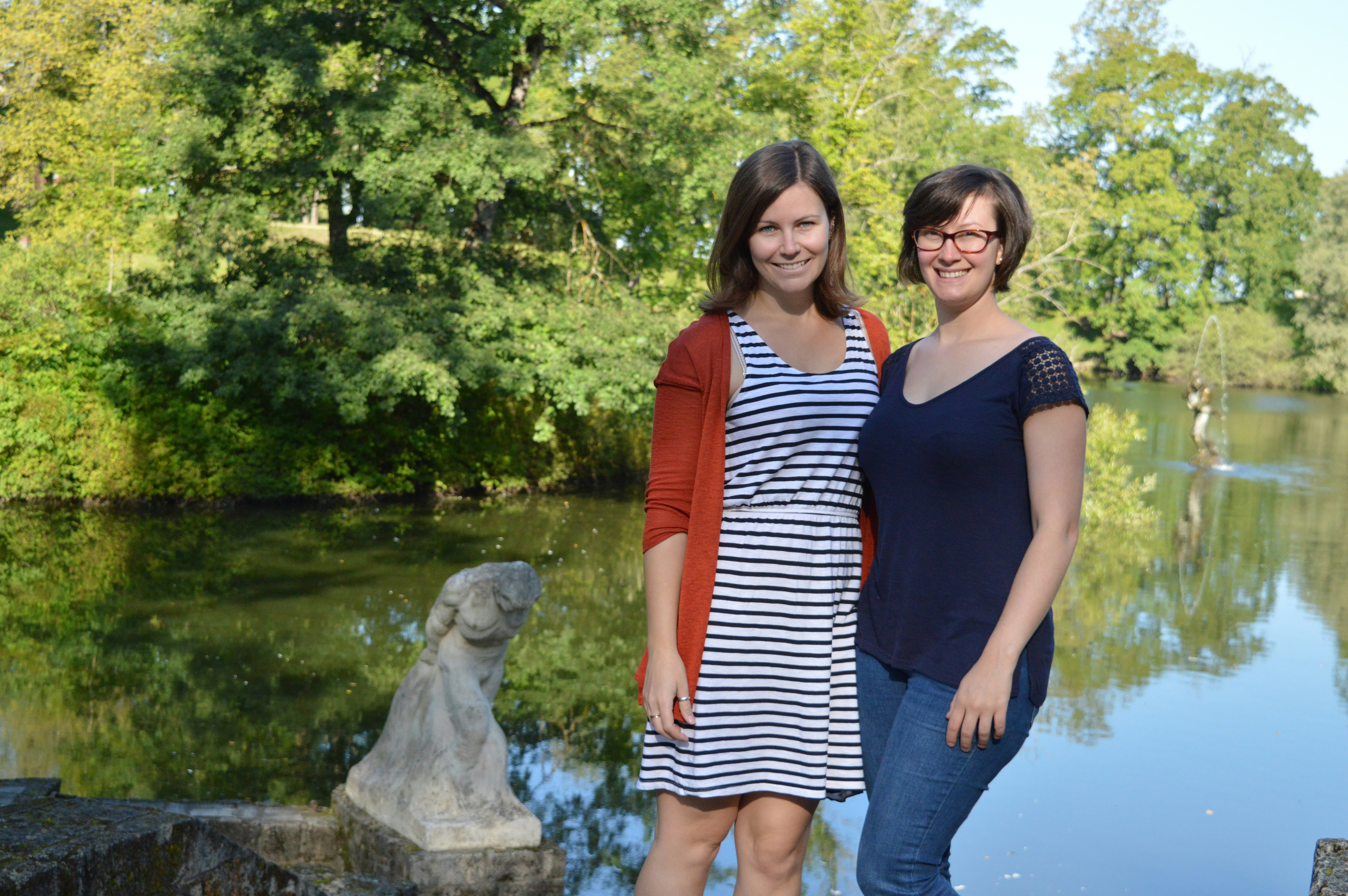 Emma and her friend Helen stand in front of a green lake with trees in the background and a statue in front of it.