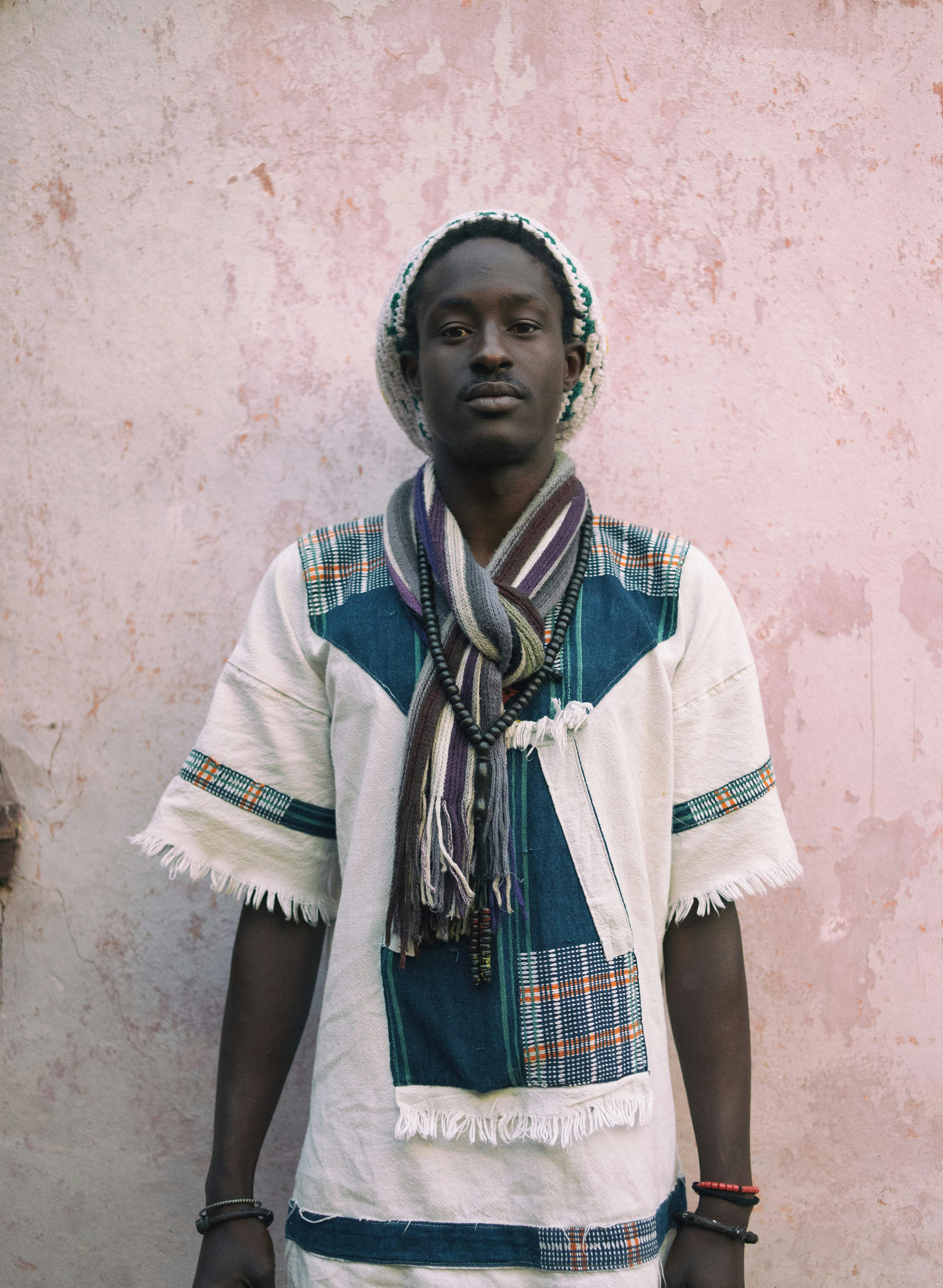 Fallou Kandji stands in front of a pink-hued wall; he's wearing a rough cotton shirt and scarf, along with a bead necklace and woven beanie.