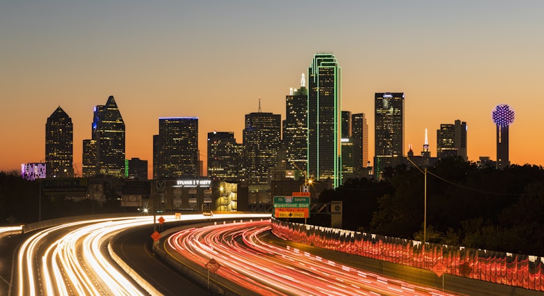 Buildings making up the downtown skyline shine bright in the night sky. Below is a blur of headlights on the highway