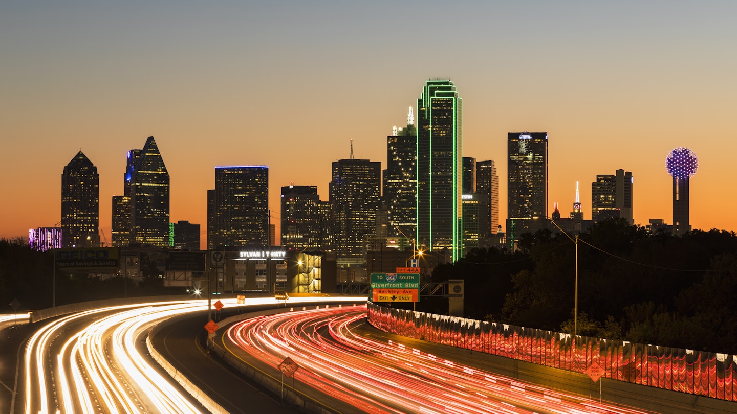 Buildings making up the downtown skyline shine bright in the night sky. Below is a blur of headlights on the highway