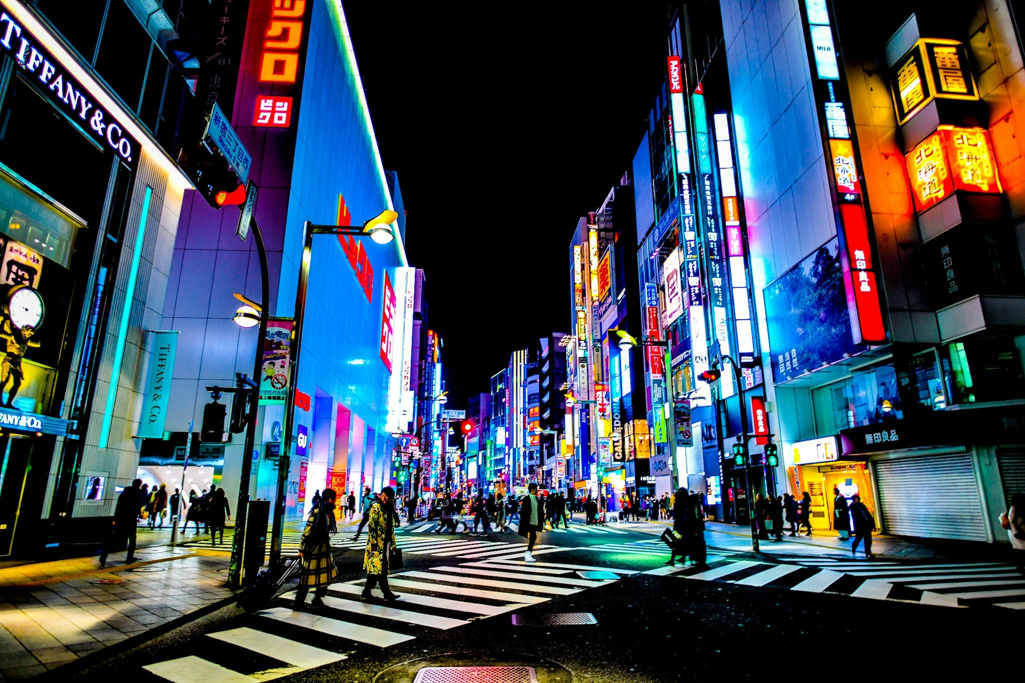 A street scene in Tokyo. Neon lighting illuminates the walls, while, below, large groups of people walk along the road.