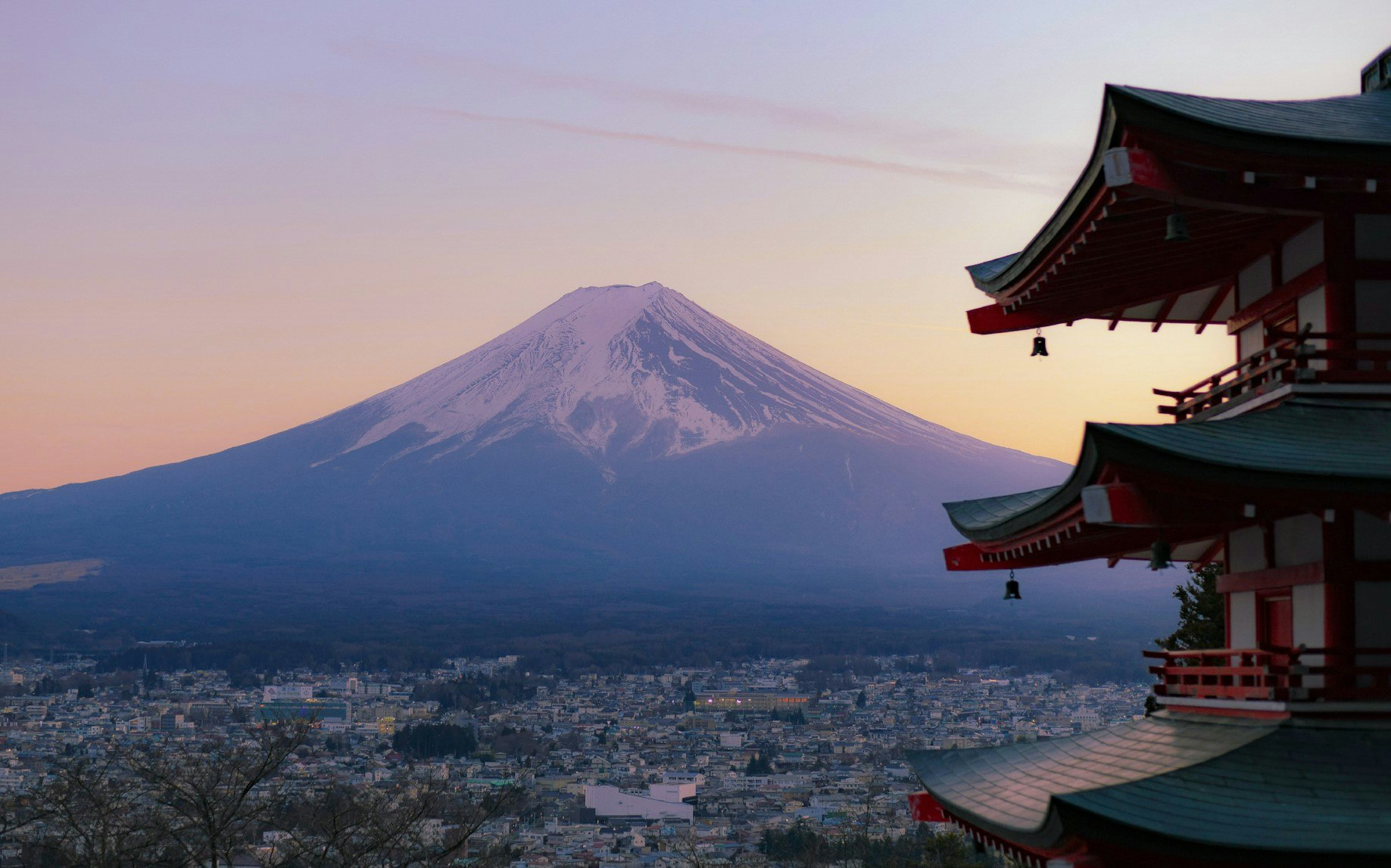 A landscape photograph of Mt Fuji from a distance. The mountain has been photographed at dusk and has a pink tint to it; a small town is visible in the shadow of the mountain. In the foreground a Japanese temple occupies the right side of the image.