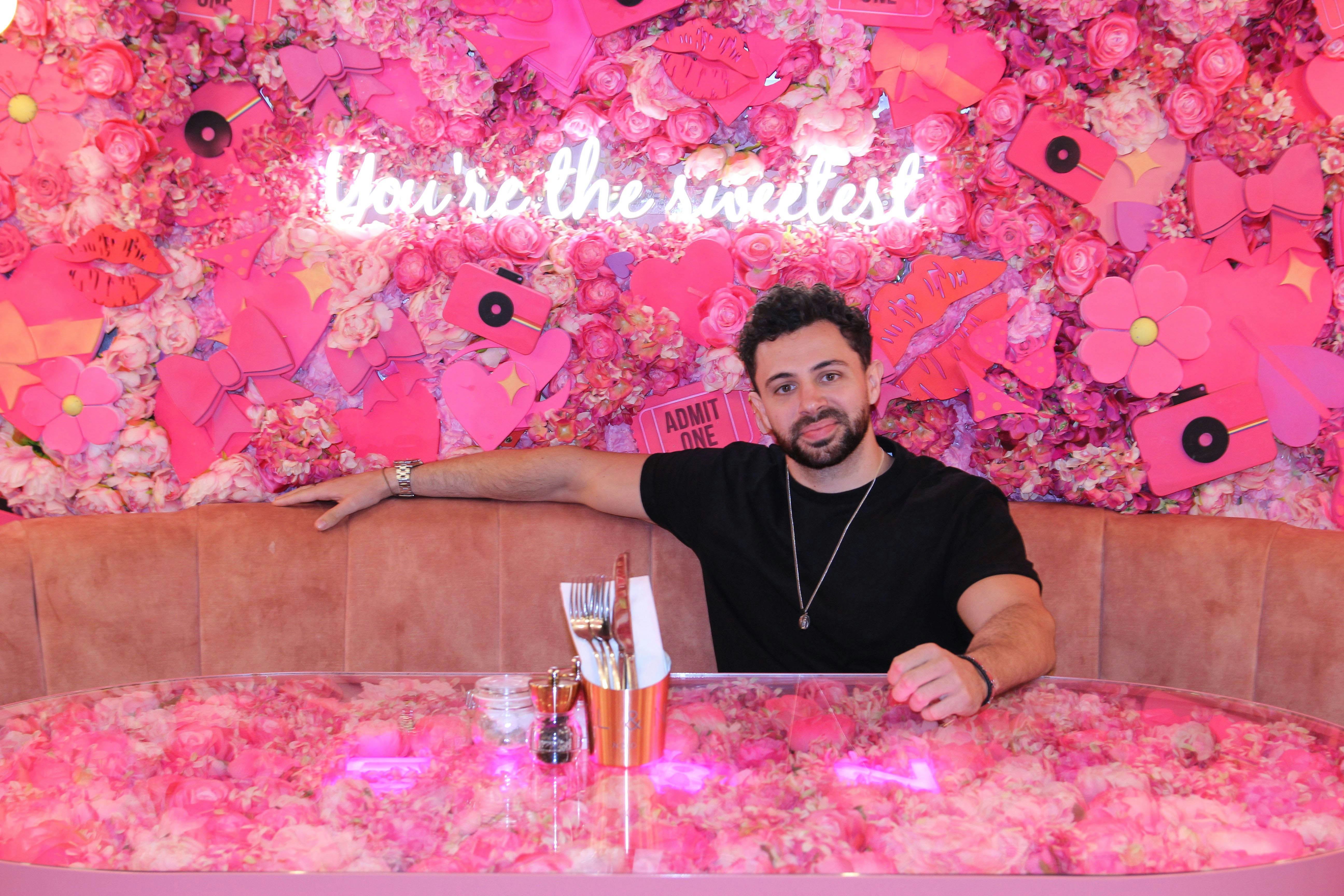 A man in a black shirt sits with his arm across the back of a booth, with a pink flowered table and matching wall with the words "You're the sweetest" written in glowing cursive