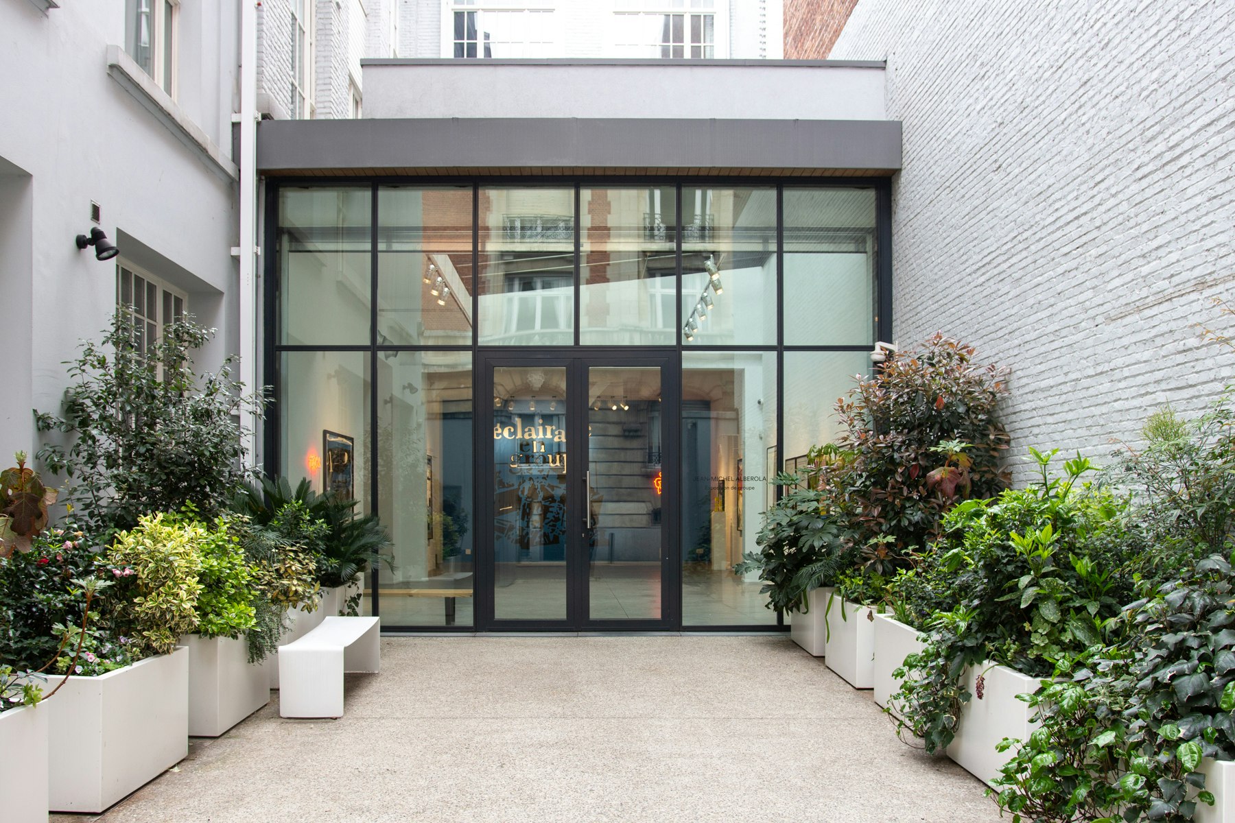 The metal and glass entrance to the Daniel Templon gallery; it's at the end of a short approach lined with greenery in planters; there are taller grey buildings on either side.