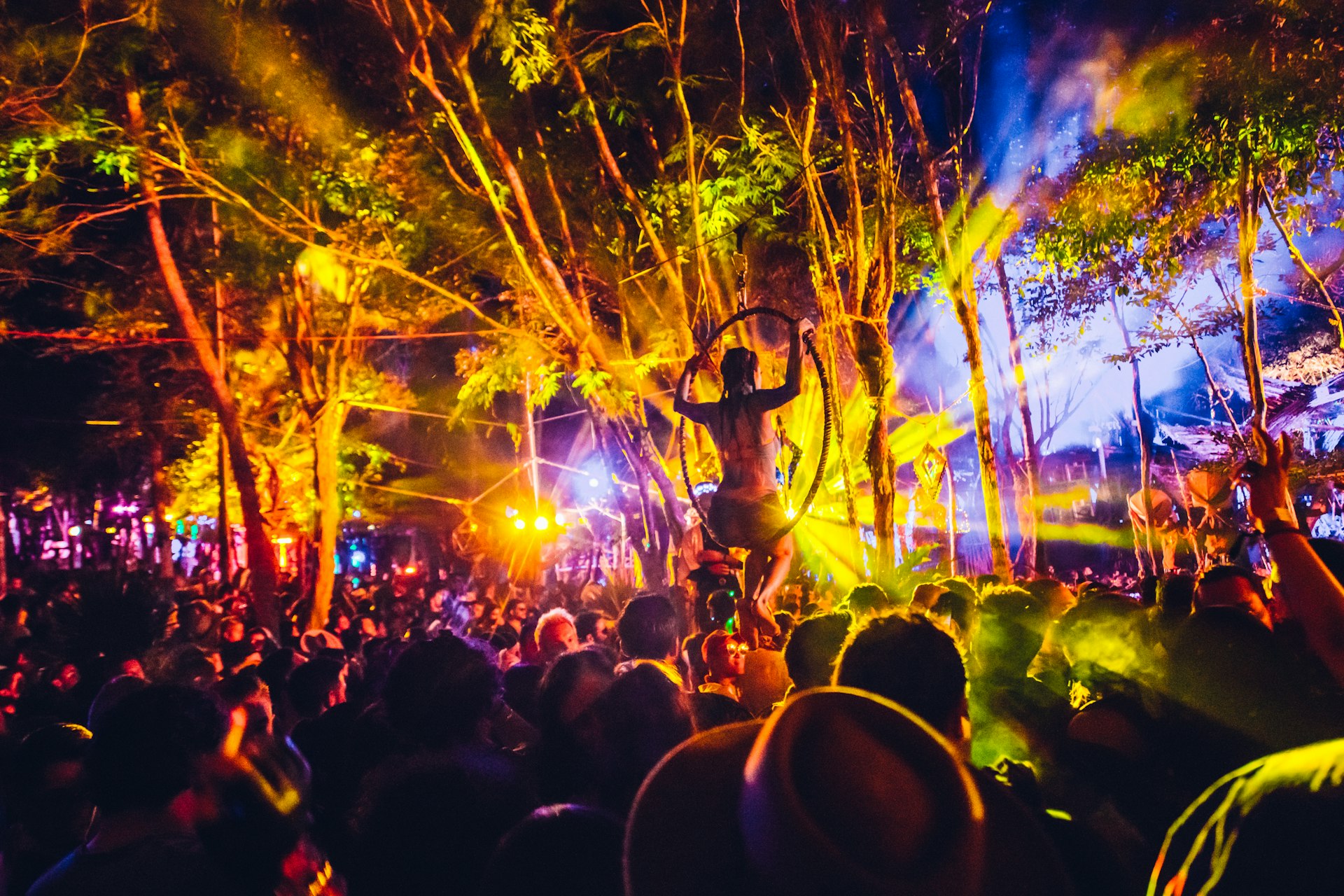 a huge crowd is gathered in front of a stage at night time. Multi-coloured lights are beaming from the stage illuminating the crowd including a performer suspended over the crowd in a hoop at Day Zero Festival in Masada, Israel. 