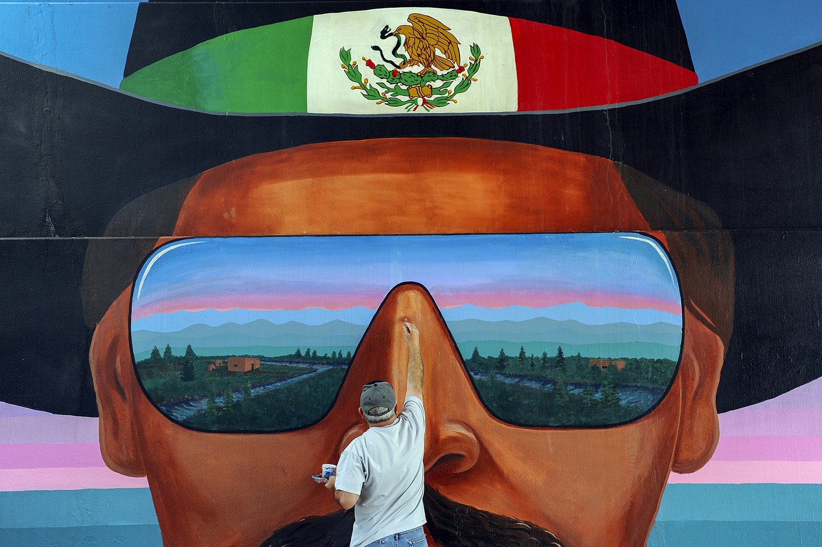 An artist touches up a mural depicting a man wearing a cowboy hat with the Mexican flag on its band and sunglasses reflecting Colorado scenery. Denver, Colorado.