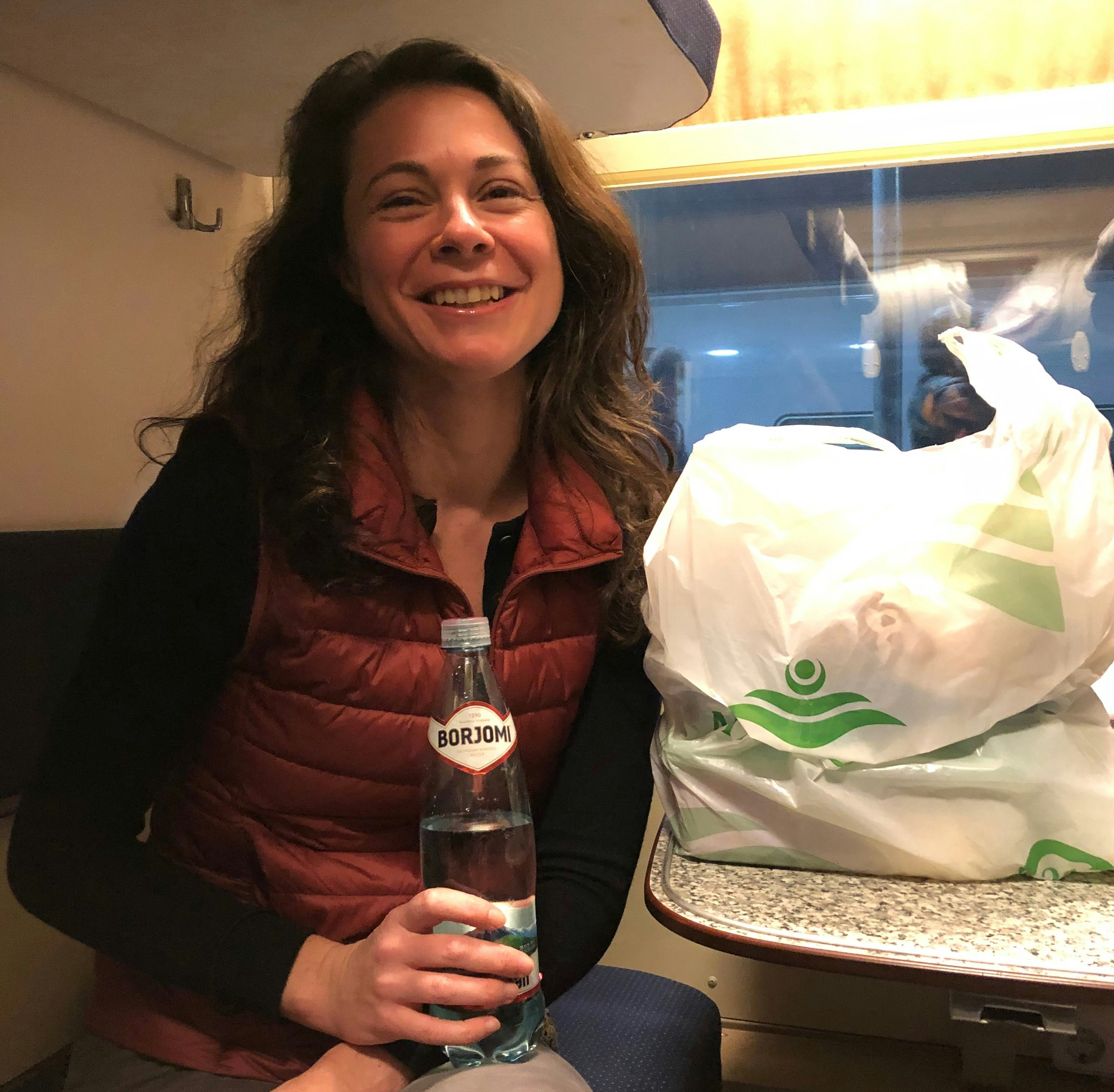 Writer Susie Armitage smiles at the camera as she sits on the bed in a small train sleeping compartment. There is a plastic bag containing groceries on a table.