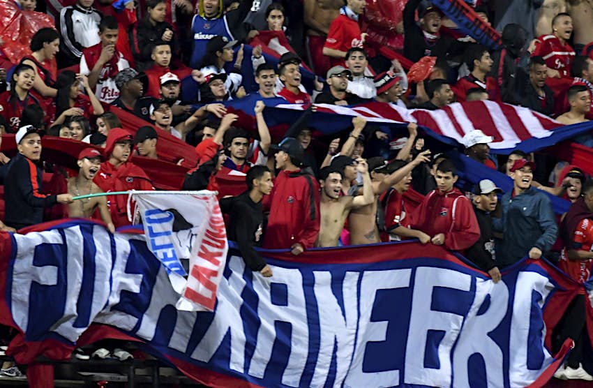 Deportivo Independiente Medellín.jpg A group of Deportivo Independiente Medellín soccer fans some wearing red and black jerseys and others shirtless wave flags and hold up large banners during a soccer game in Colombia.
