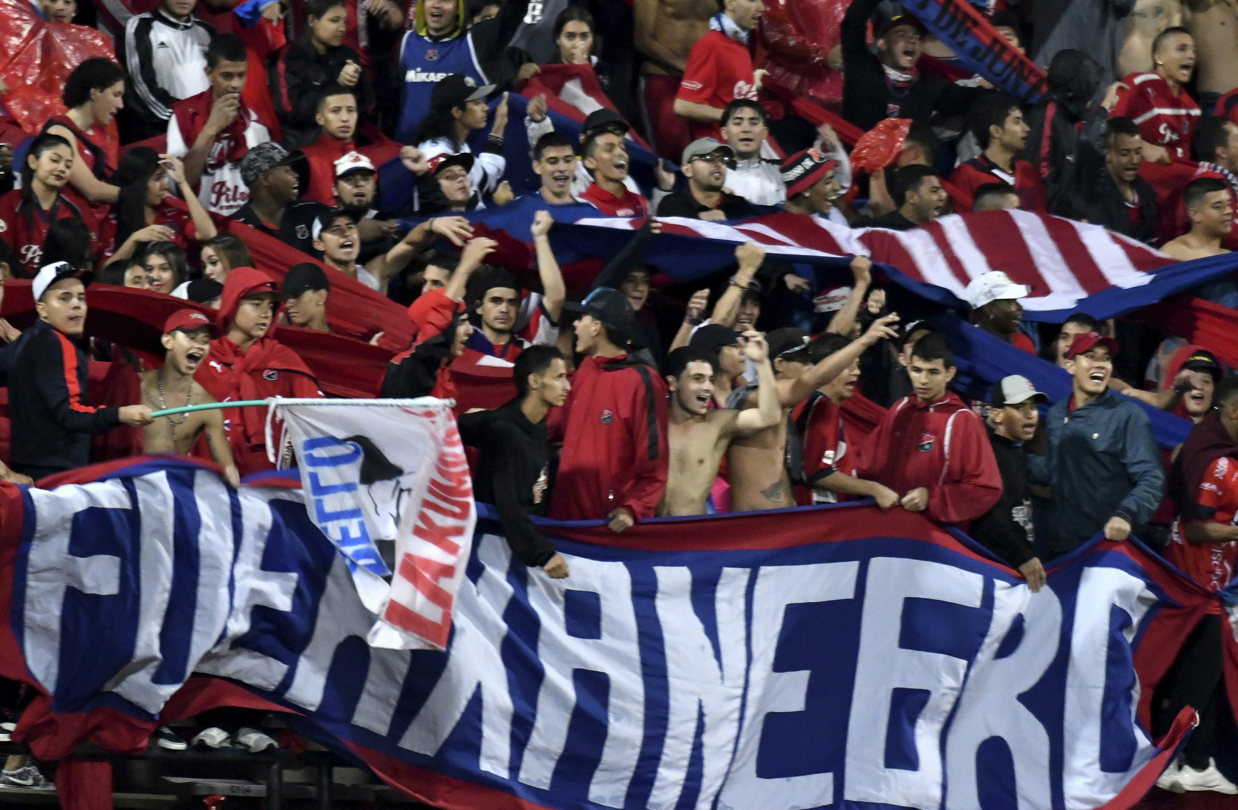 A group of Deportivo Independiente Medellín soccer fans some wearing red and black jerseys and others shirtless wave flags and hold up large banners during a soccer game in Colombia.