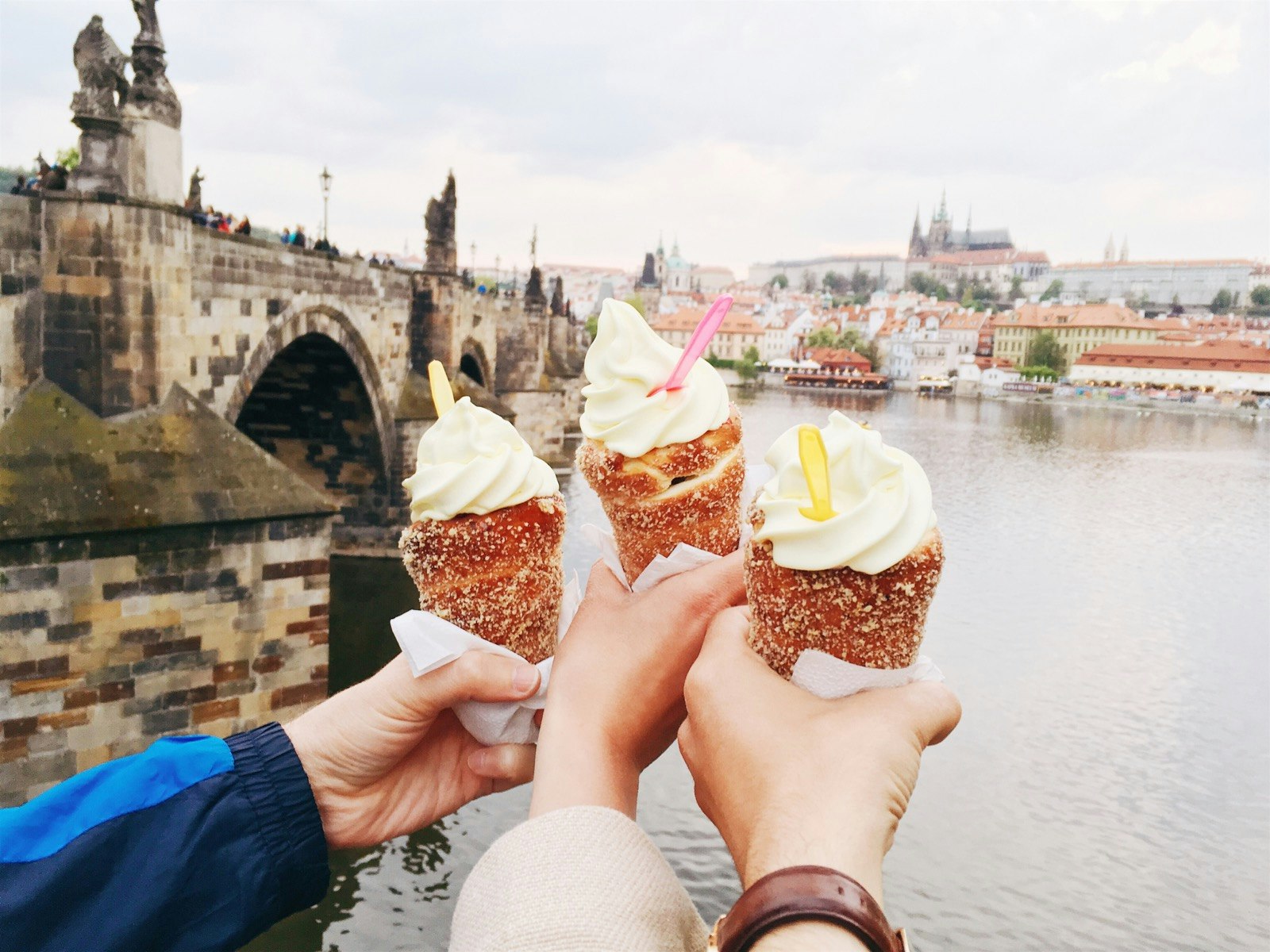 Three hands hold cinnamon rolls topped with ice cream with the Prague skyline in the backgroudn