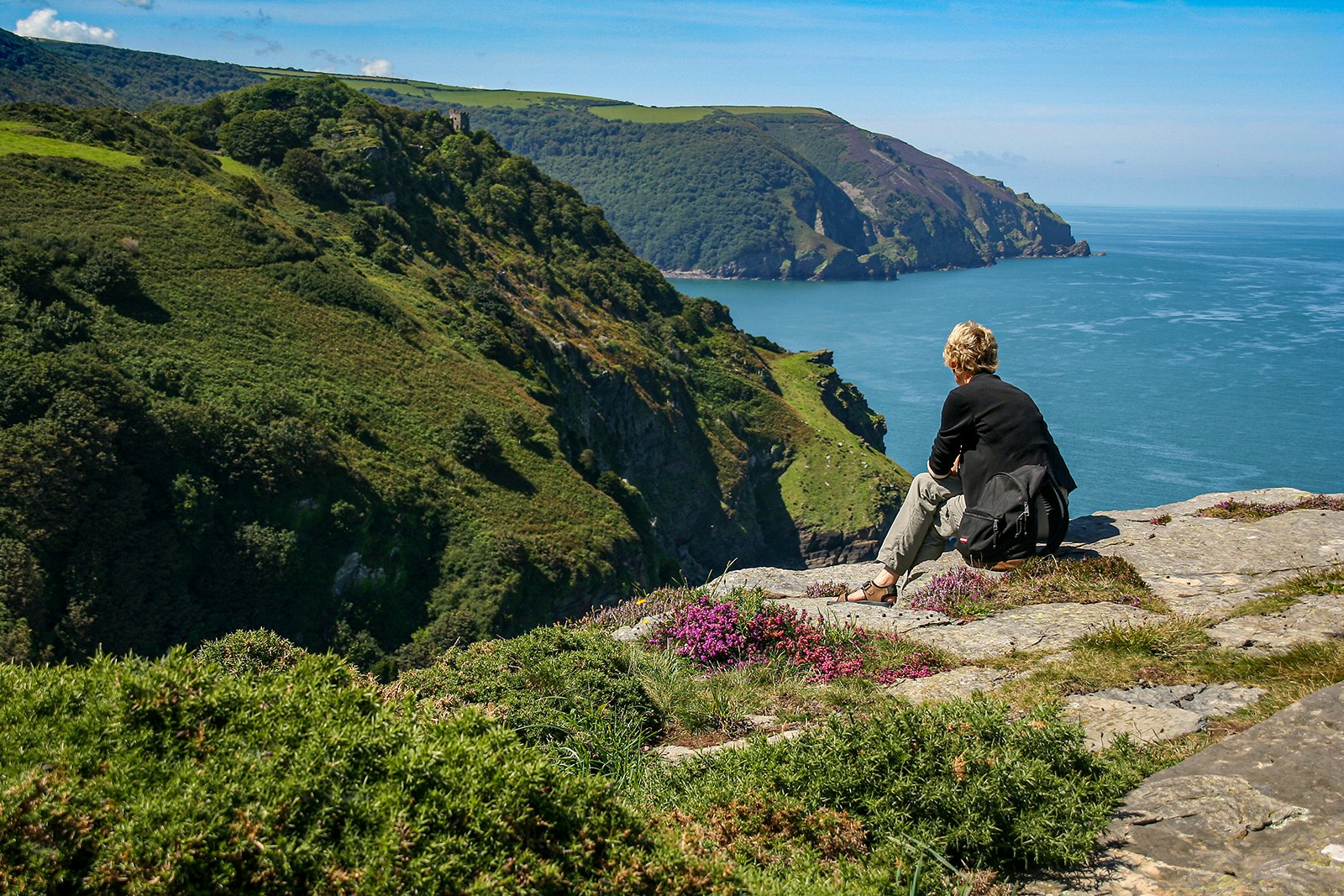 A blond person sits on a rocky outcropping, overlooking a coastline abutted by green cliffs. England.