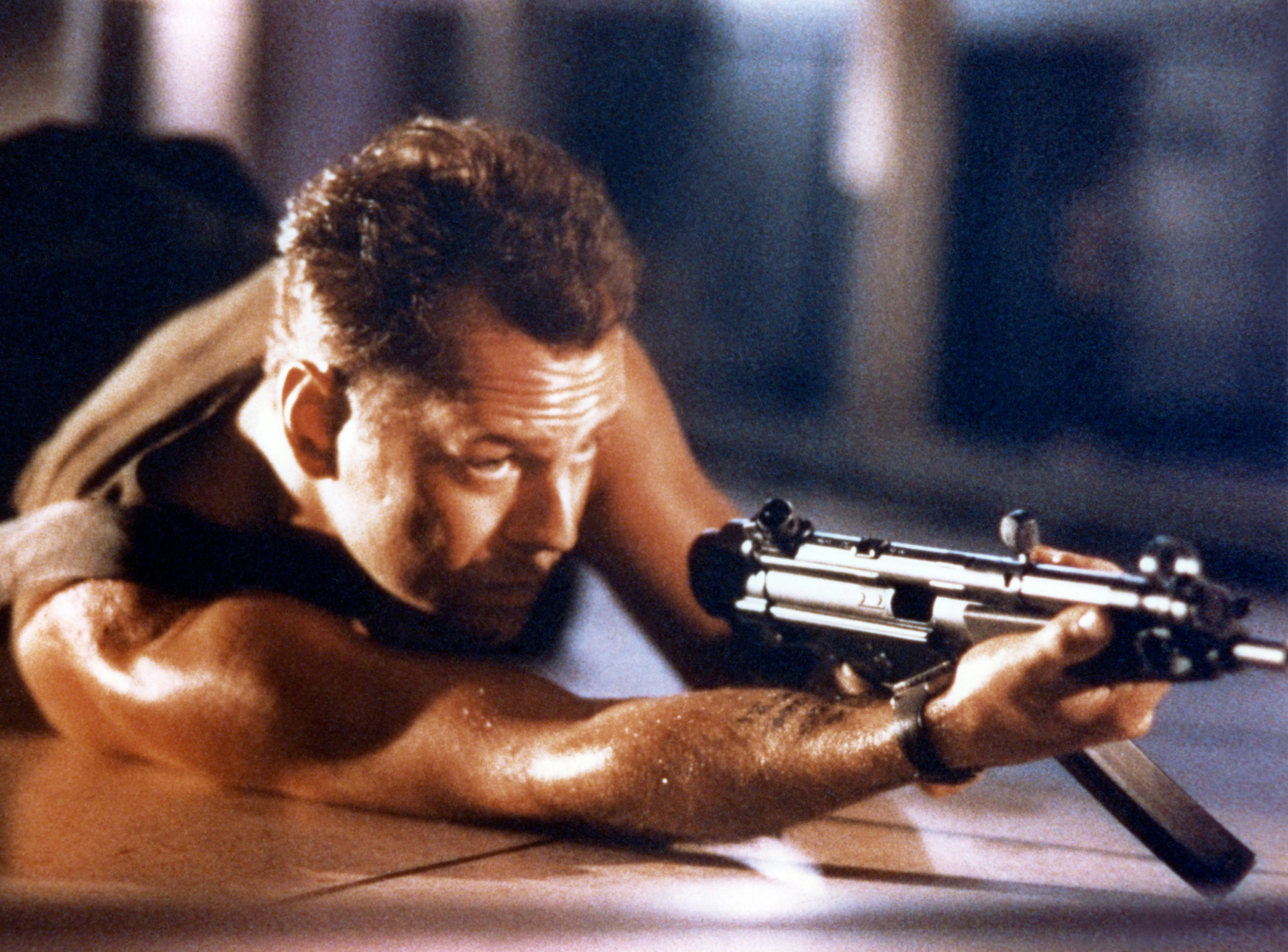 A sweaty and vested Bruce Willis aims a machine gun in Die Hard