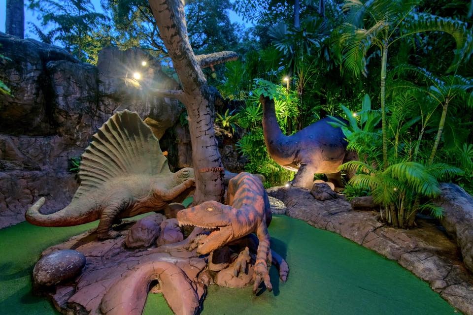 Large dinosaur models are around a miniature golf course in Phuket, Thailand. A floodlight is illuminating the course.