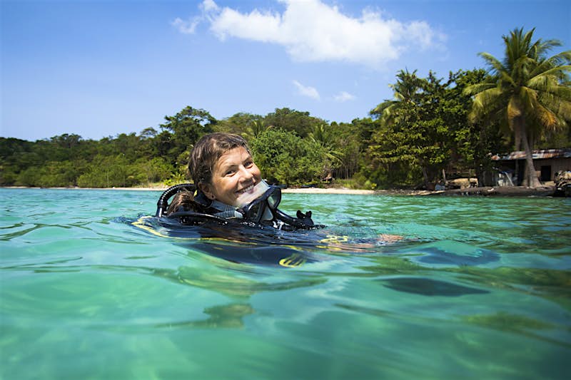 The head of a female dive instructor pokes out of the azure waters; her mask is around her neck, and the rest of her equipment under the water. The shore in the background is lined with palms.