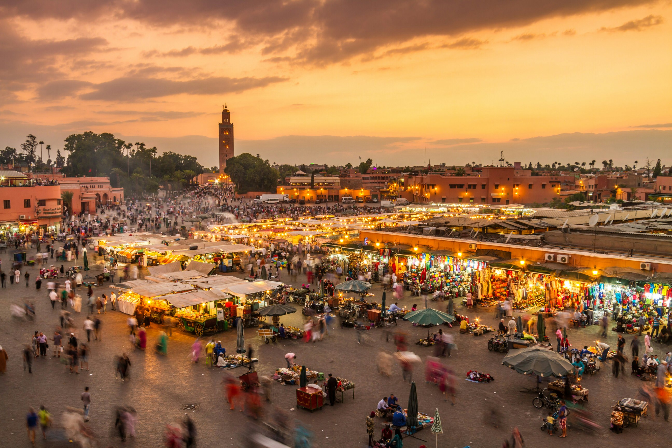 Aerial of a crowd at Djemaa El Fna square during the early evening in Marrakesh's Medina quarter; moving people are blurred between the umbrella-covered food stands. The mosque's minaret stands tall against the orange sky in the background.
