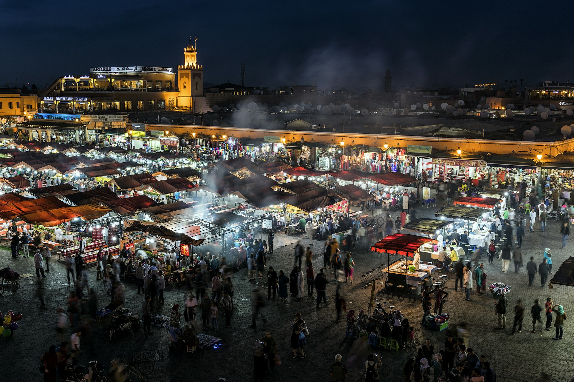 A market square at night with people milling around food stalls cooking fresh dishes