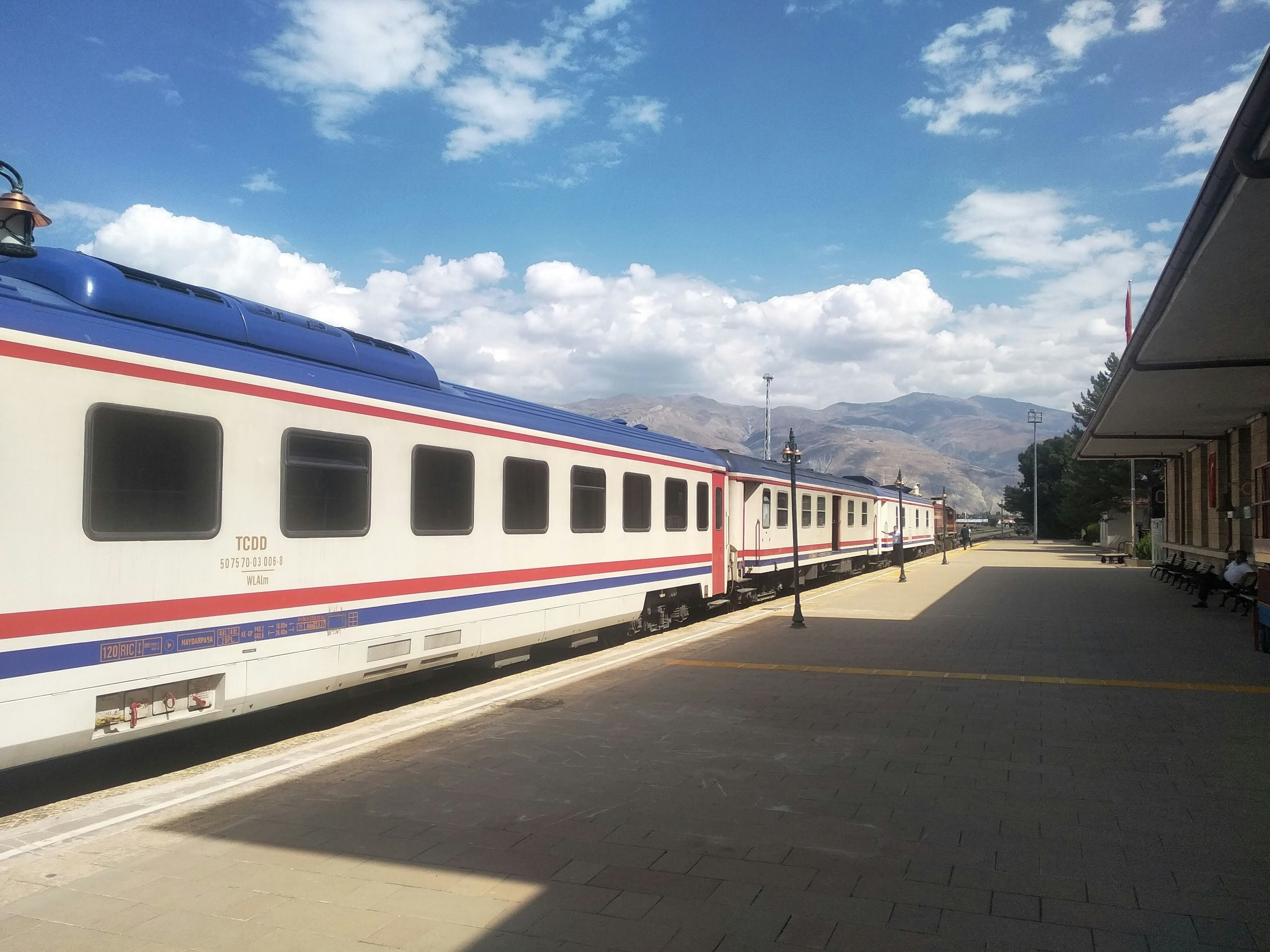 Three carriages of a train standing at a station platform. Each carriage is white with two stripes in red and blue running the length of the carriage below the windows. There are hills in the background