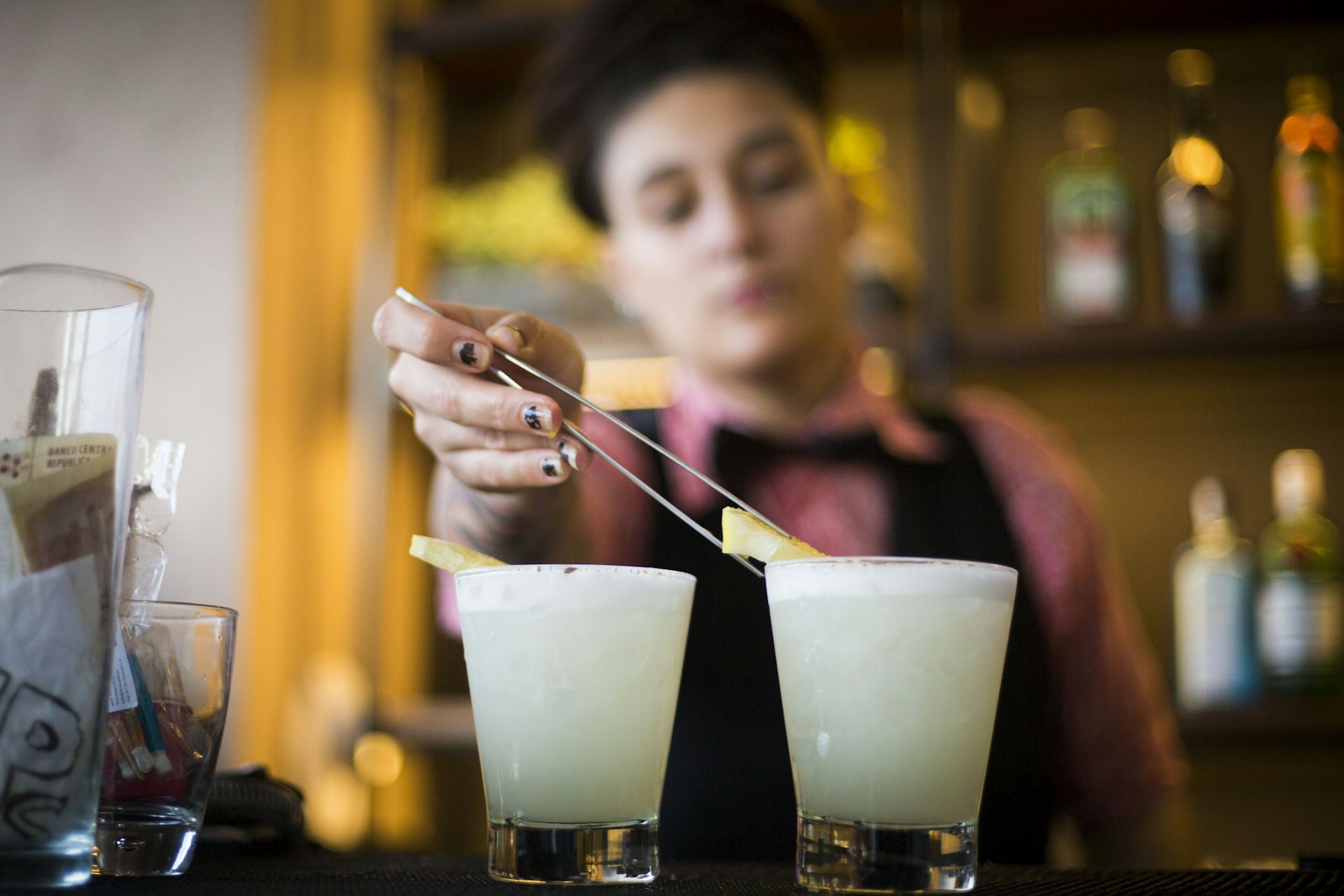 A bartender, holding a large tweezer places a piece of fruit into a lightly-colored drink at the bar; Buenos Aires rooftop bars   