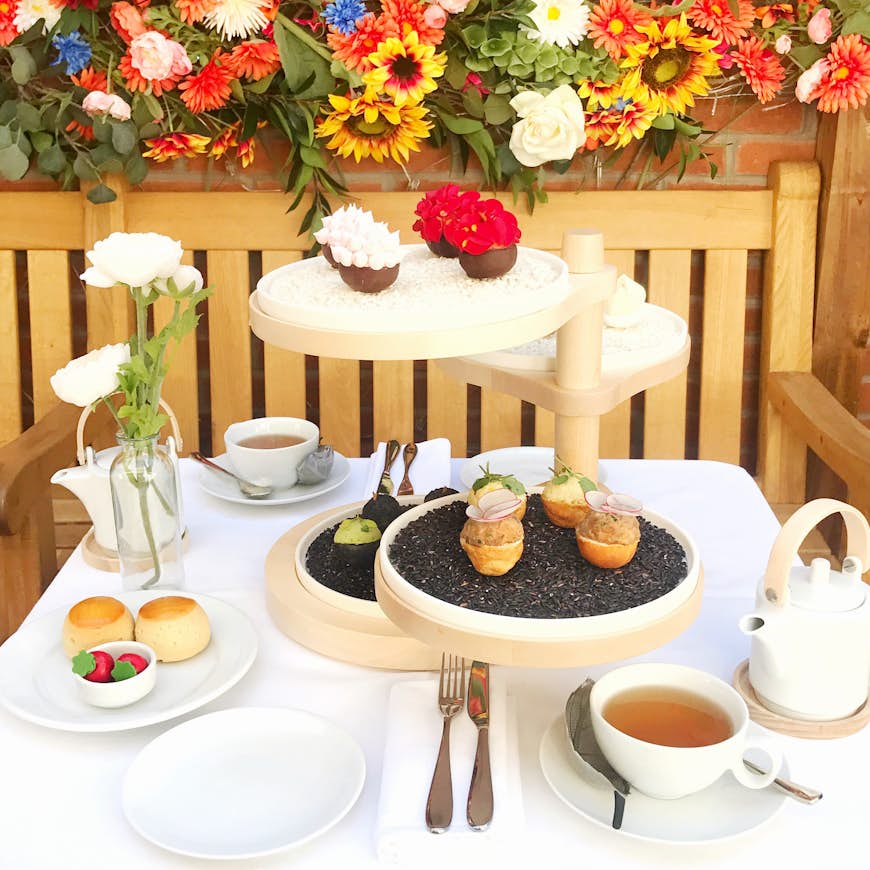 White cups of tea on a white tablecloth with pastries on a wooden stand; there are bright colourful flowers above a wooden bench in the background.
