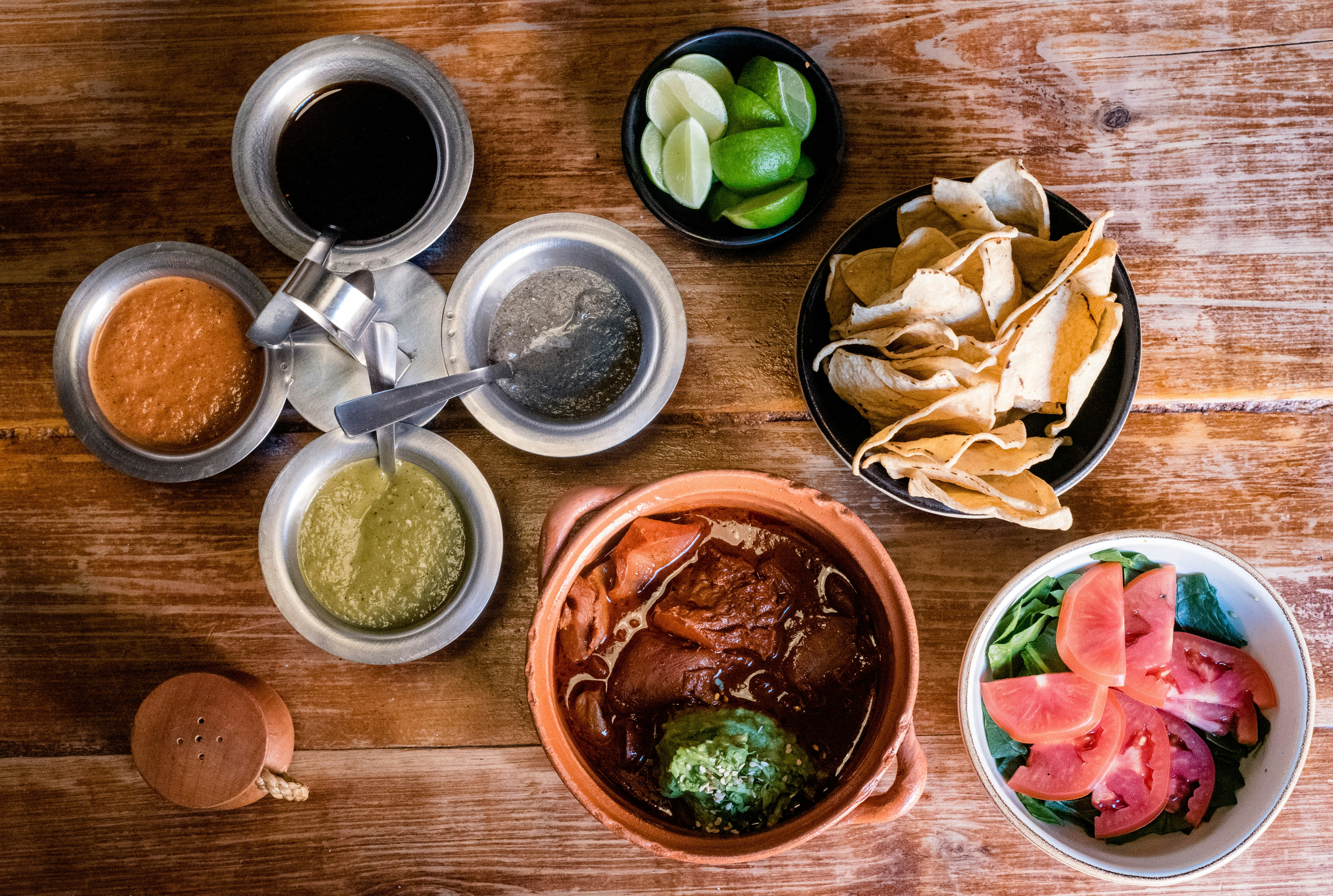 Aerial view of tacos, sliced limes and tomatoes, and various sauces, arranged on a wooden table