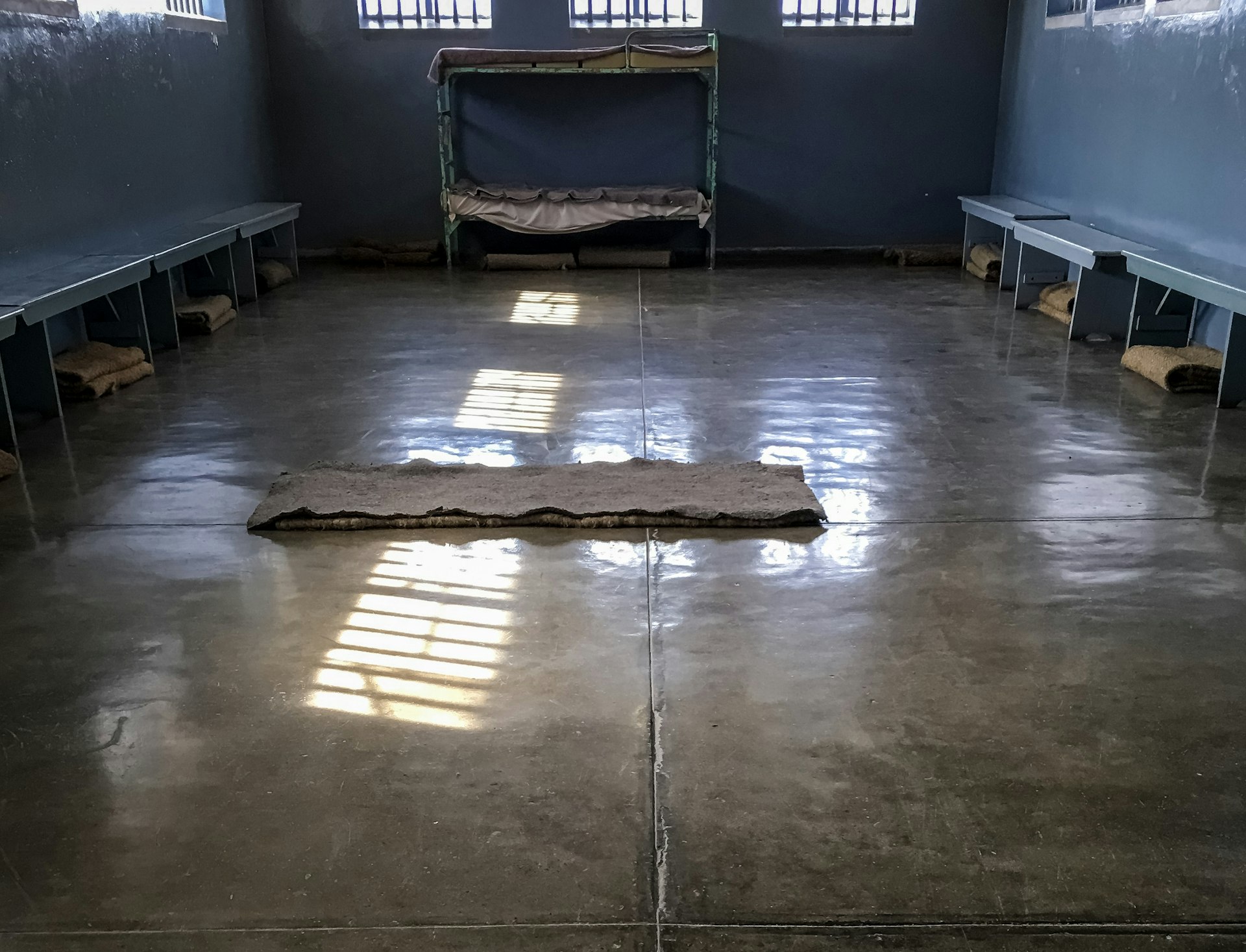 A sisal mat with rough blanket lay atop a polished concrete floor within a dark grey room with barred widows; benches line the walls and a single bunk bed sits at the far end.