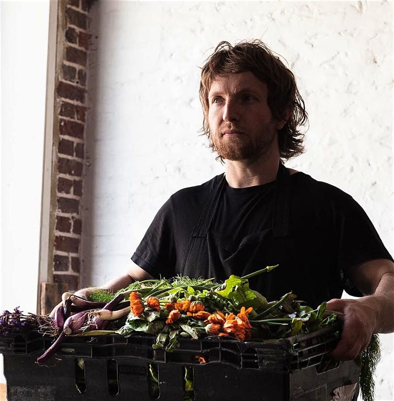 A man in black holds a basket full of vegetables in front of a white background. 