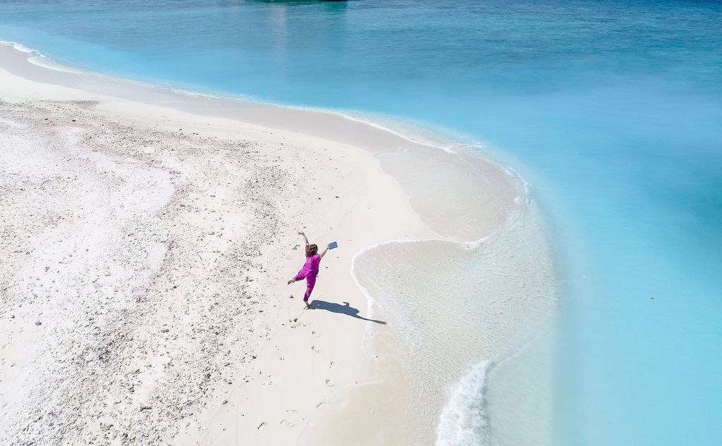 Drone shot of woman walking on a white sand beach with a book in hand