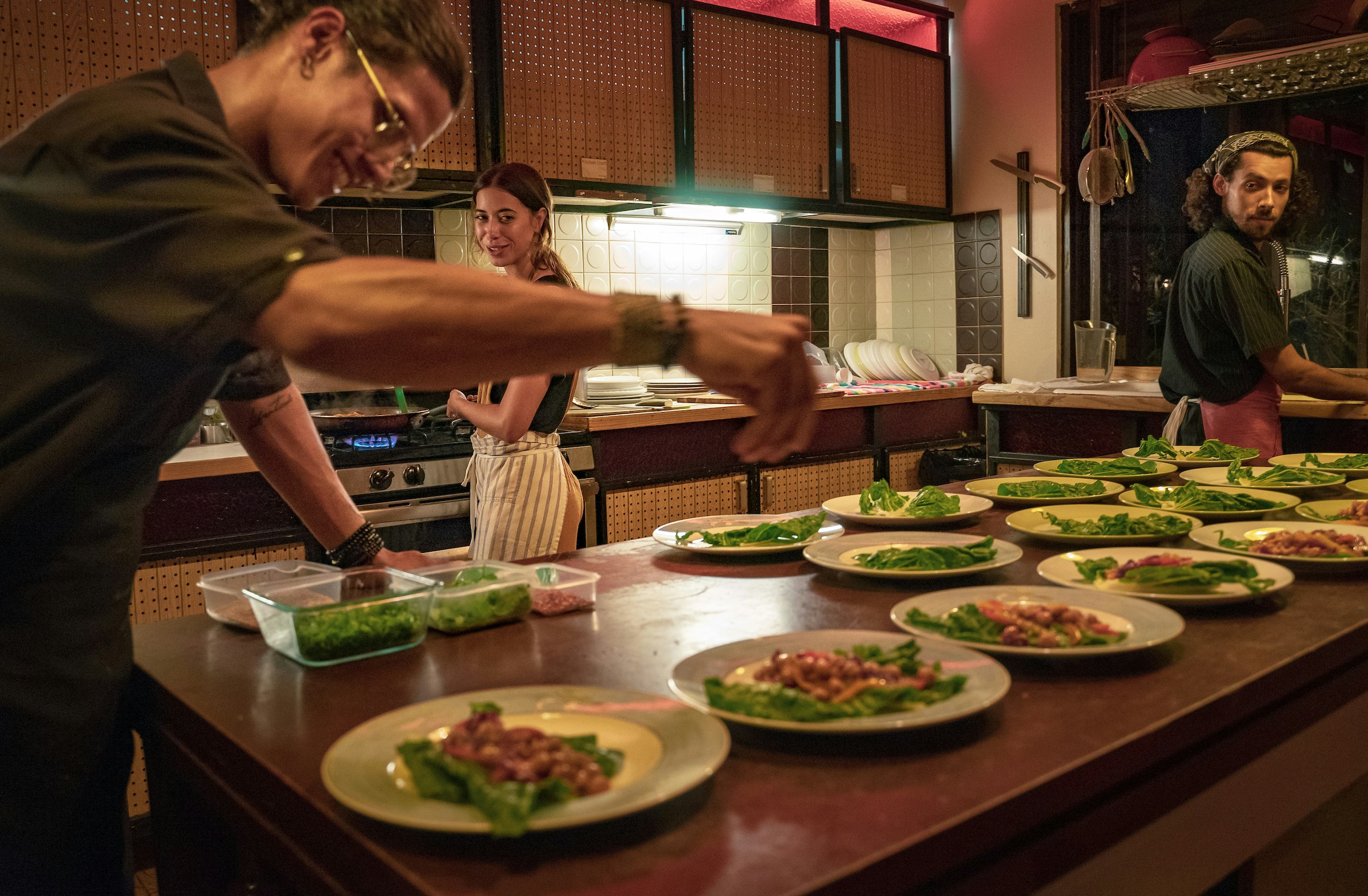 A chef sprinkles garnish over a plate of food as a pair of other chefs look on. The table is filled with dishes filled with food in a kitchen; Sustainable Puerto Rico 