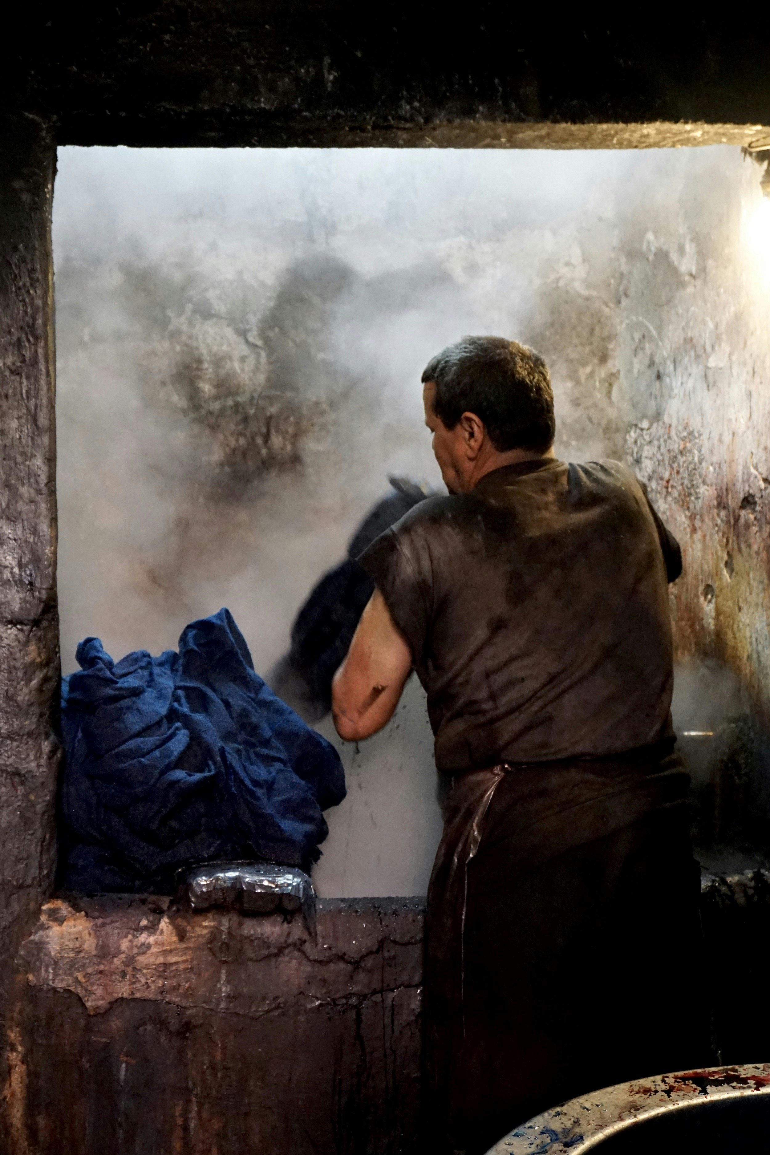 A worker, with his back to the camera, pulls wet fabrics from a steaming pit of dye.