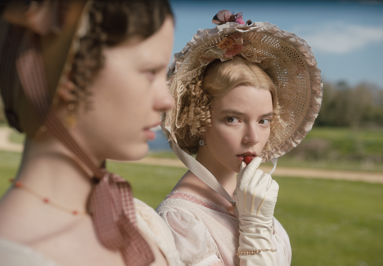 Anya Taylor-Joy as Emma in the film adaptation of Jane Austen's novel. She is looking over her right shoulder (while eating a strawberry) towards a woman who is out of focus. 