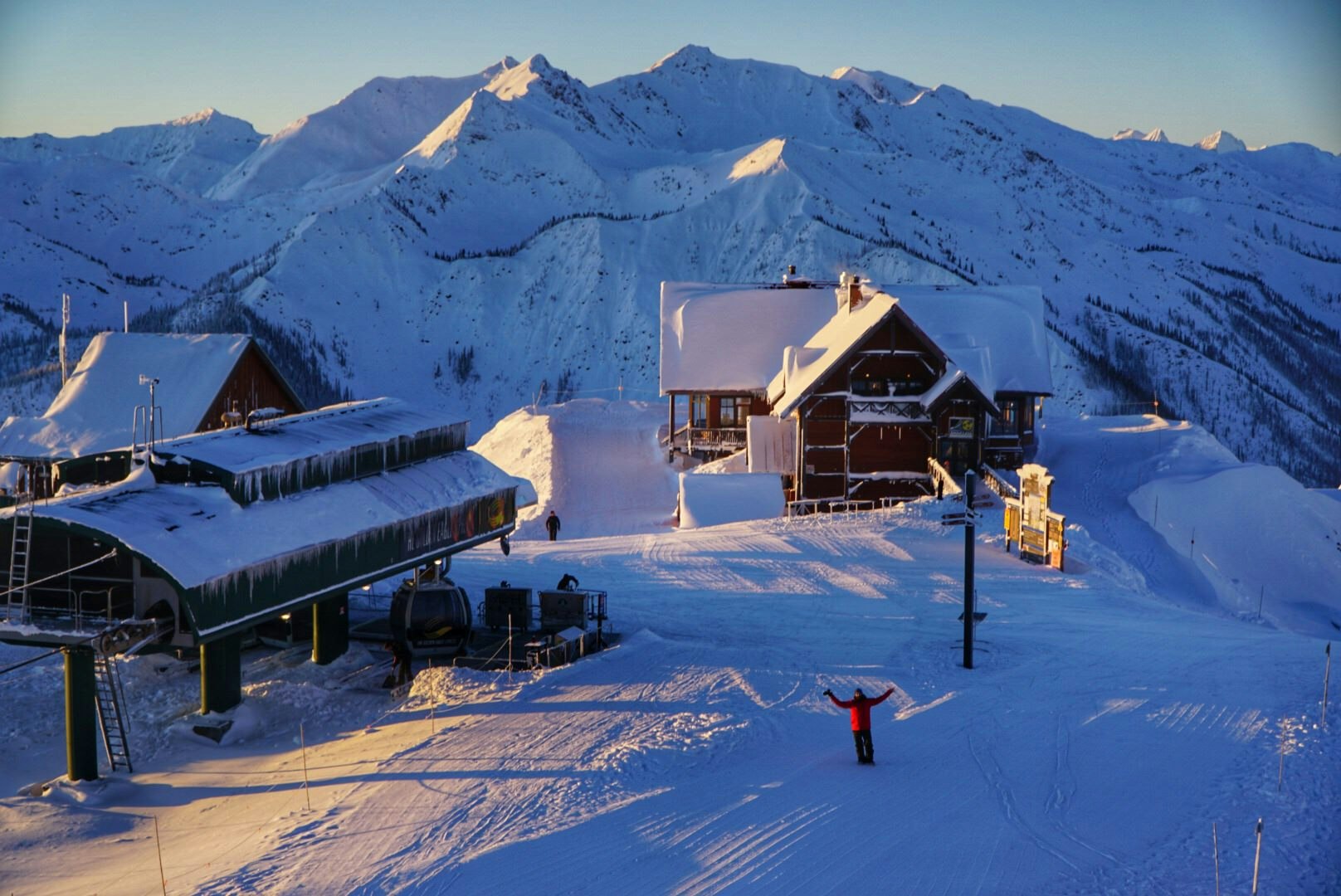 A person in a red ski jacket and black pants is joyfully holding up their arms; they're standing at the top of a piste, beside a ski-lift station. Behind them is a mountain chalet and, beyond that, are mountains covered in snow.