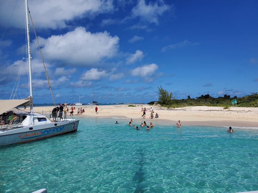 A group of people relax in bright blue ocean water, while others lounge on the sand. There is a catamaran moored in the shallow water. 