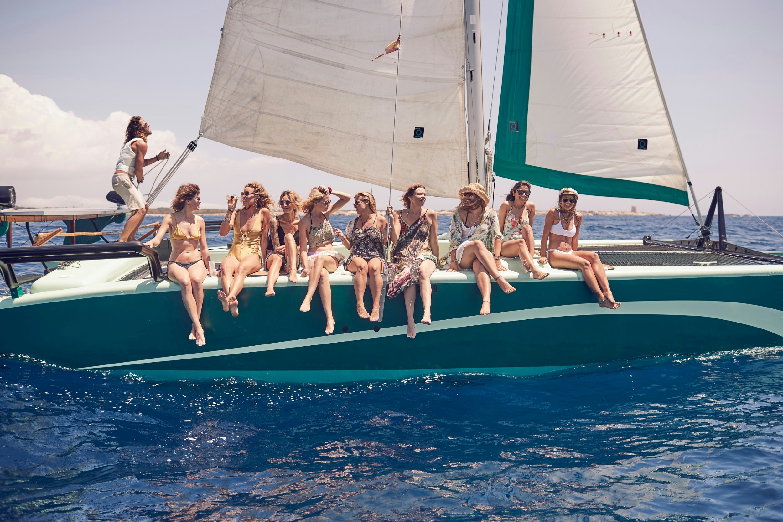 Nine women sitting on the side of a catamaran while one man sails it