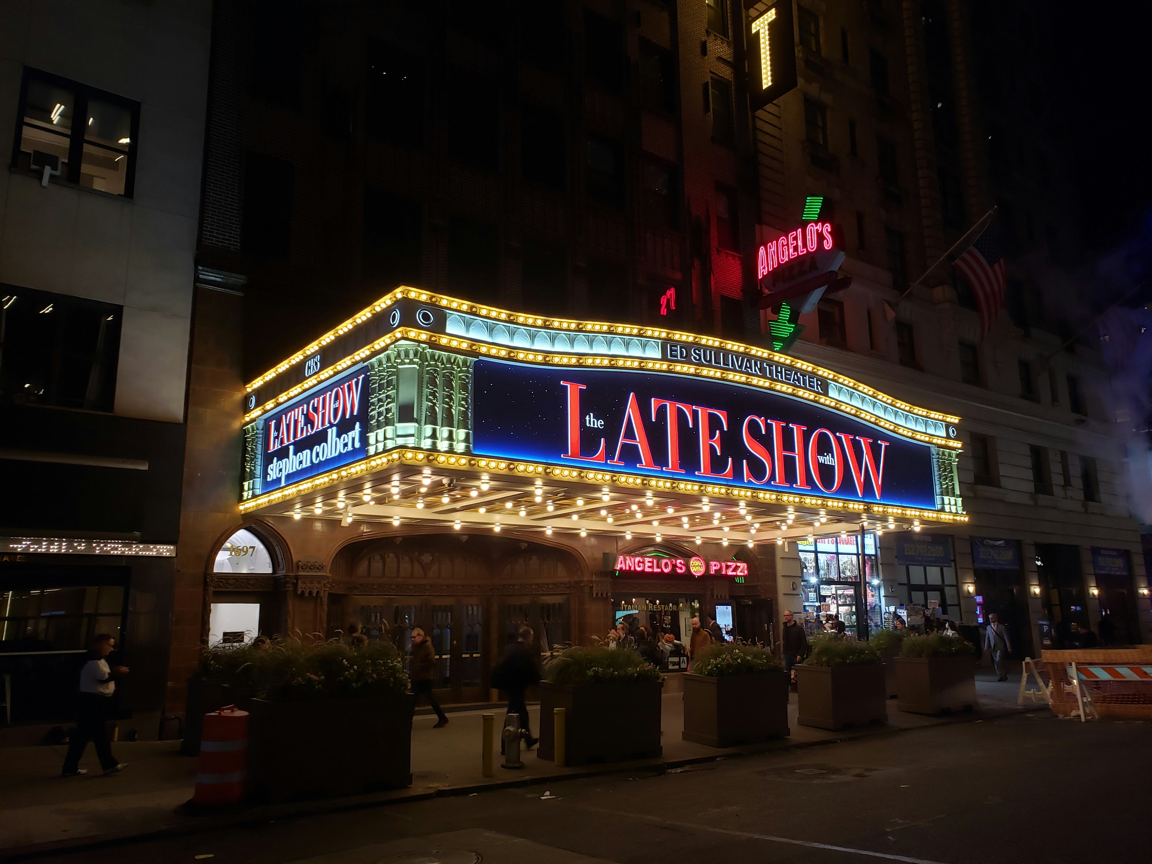 The exterior of the Ed Sullivan Theater shows a vast overhanging marquee with large-bulb theater lights on the underside and The Late Show lit up in red serif letters on the front. Large square planters that double as traffic bollards sit between the street and the entrance, where pedestrians stroll past a neon sign for Angelo's Pizza inside the theater.
