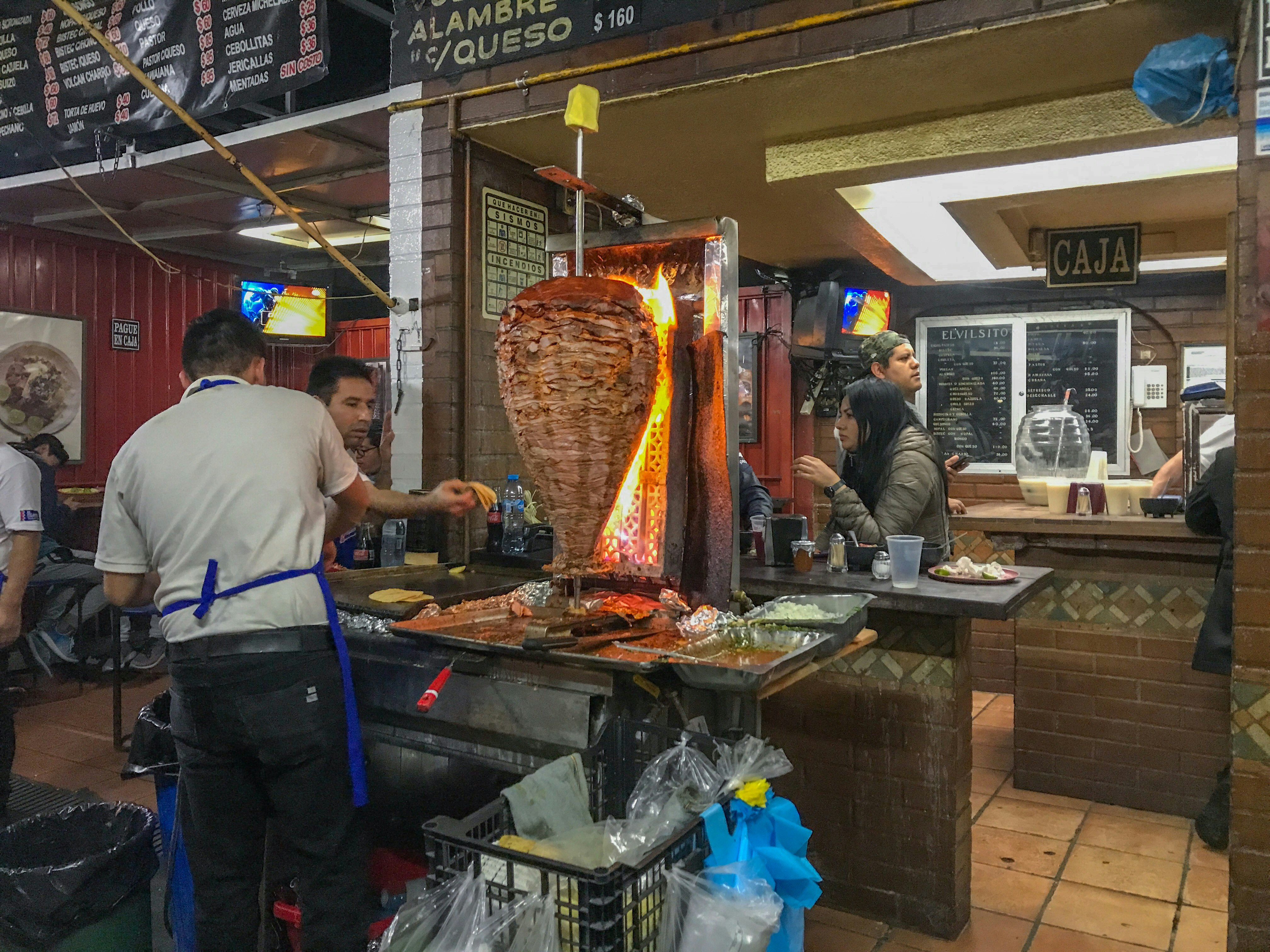 A large chunk of meat is being roasted on a large spit as a man prepares taco shells on a large grill as another man looks on. There are a group of people eating in the background. 