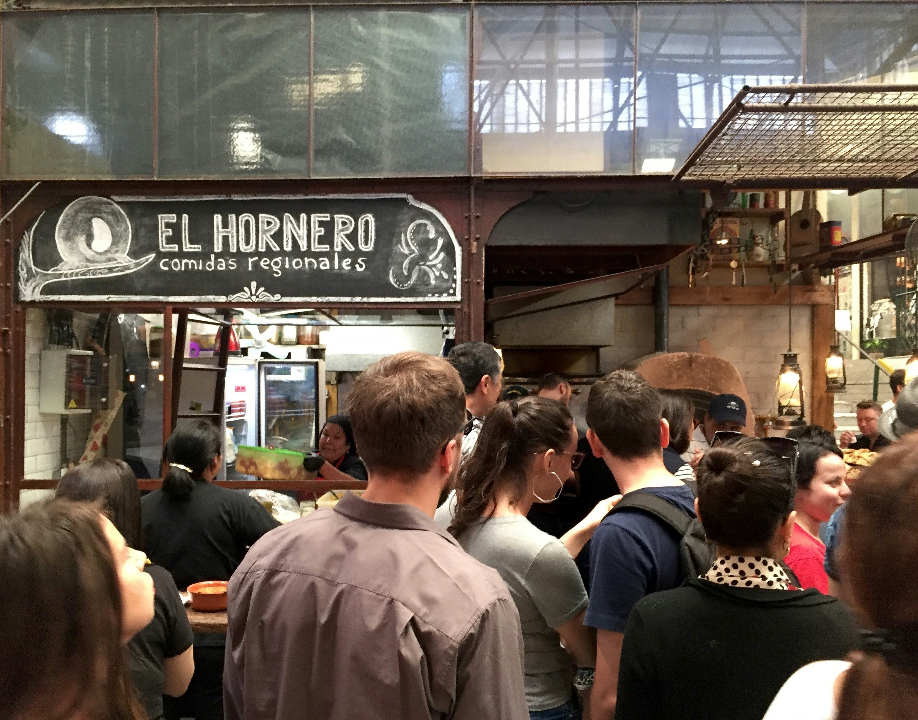 A crowd of people are lining up for empanadas at the El Hornero stall in a covered market. The stall's sign has been drawn on a chalkboard.