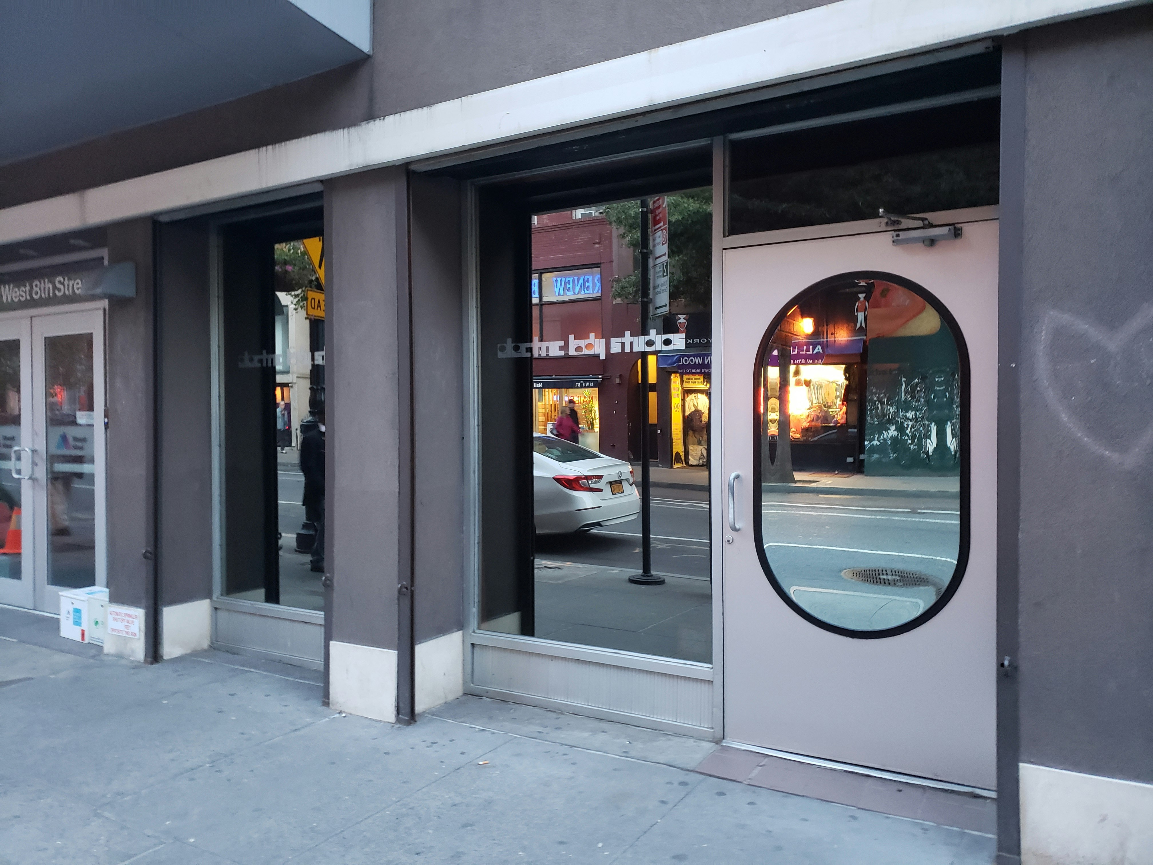 The Electric Lady Studios exterior entrance is quite unassuming, with a swinging door with an ovular, mirror-reflective window in a light purple tone with a sign reading "Electric Lady Studios" in a futuristic font