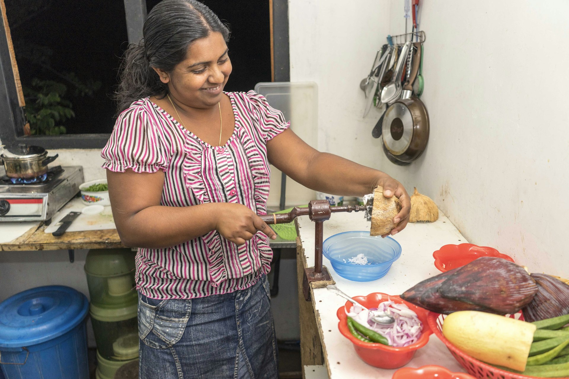 A woman is smiling while using a hand turned implement to shave the inside of a coconut. She is standing in a kitchen with lots of fresh produce on the counters and a small stove in the background. 