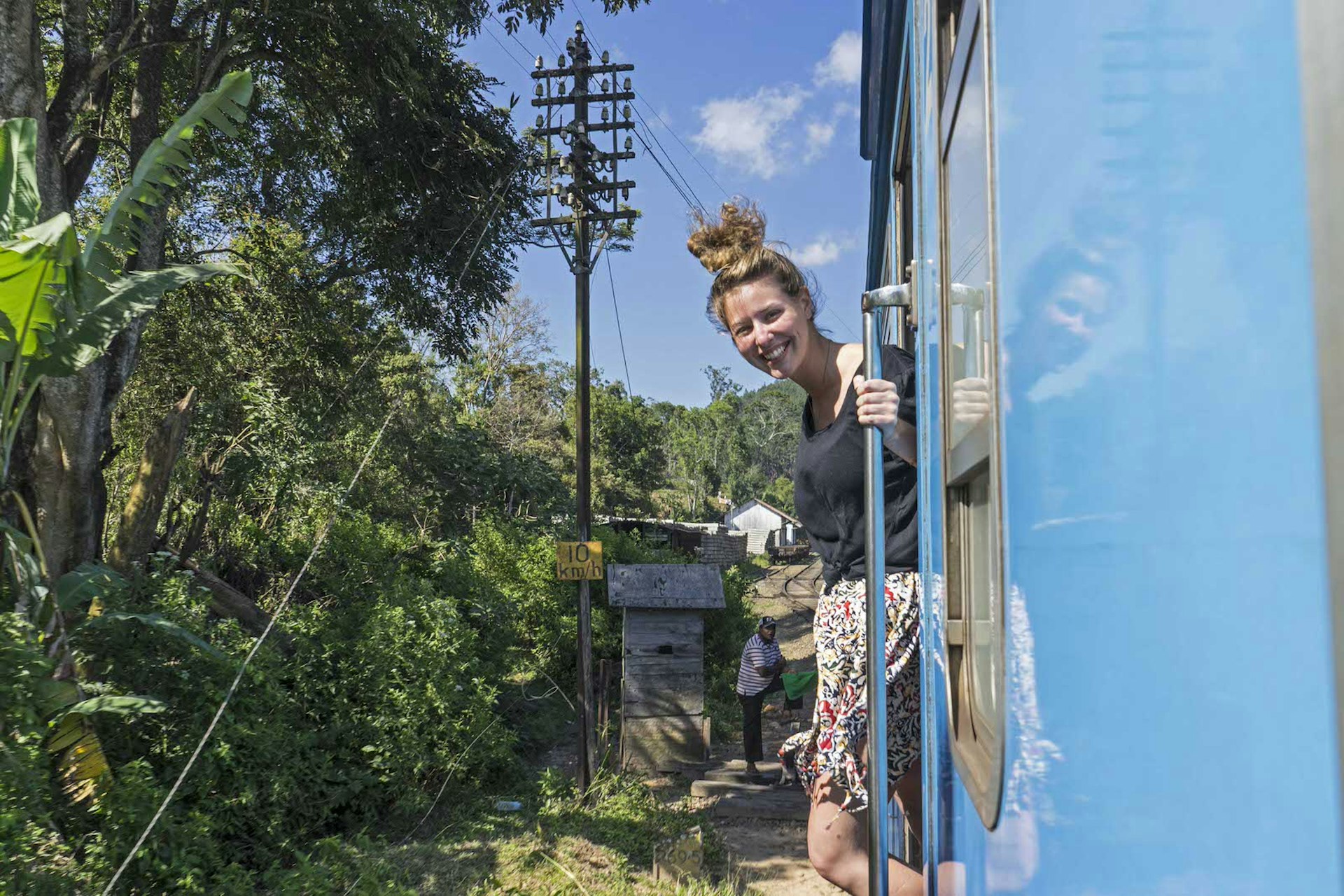 A woman hangs out of a train door smiling at the camera, positioned out of a train window. The train is bright blue and it is a sunny day in Ella, Sri Lanka. There are tropical trees next to the tracks. 