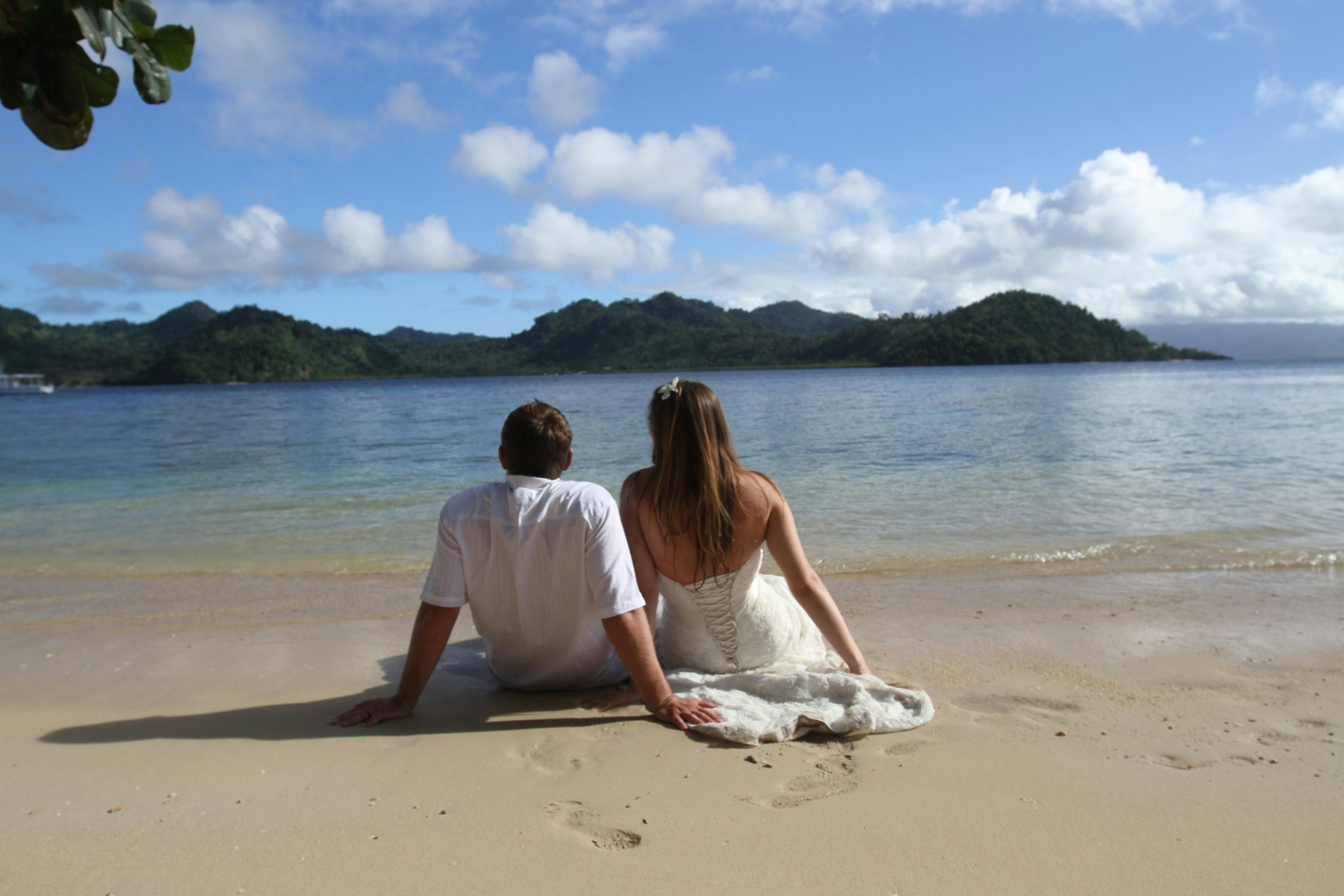 A young newlywed couple sitting on the sand looking at the ocean.